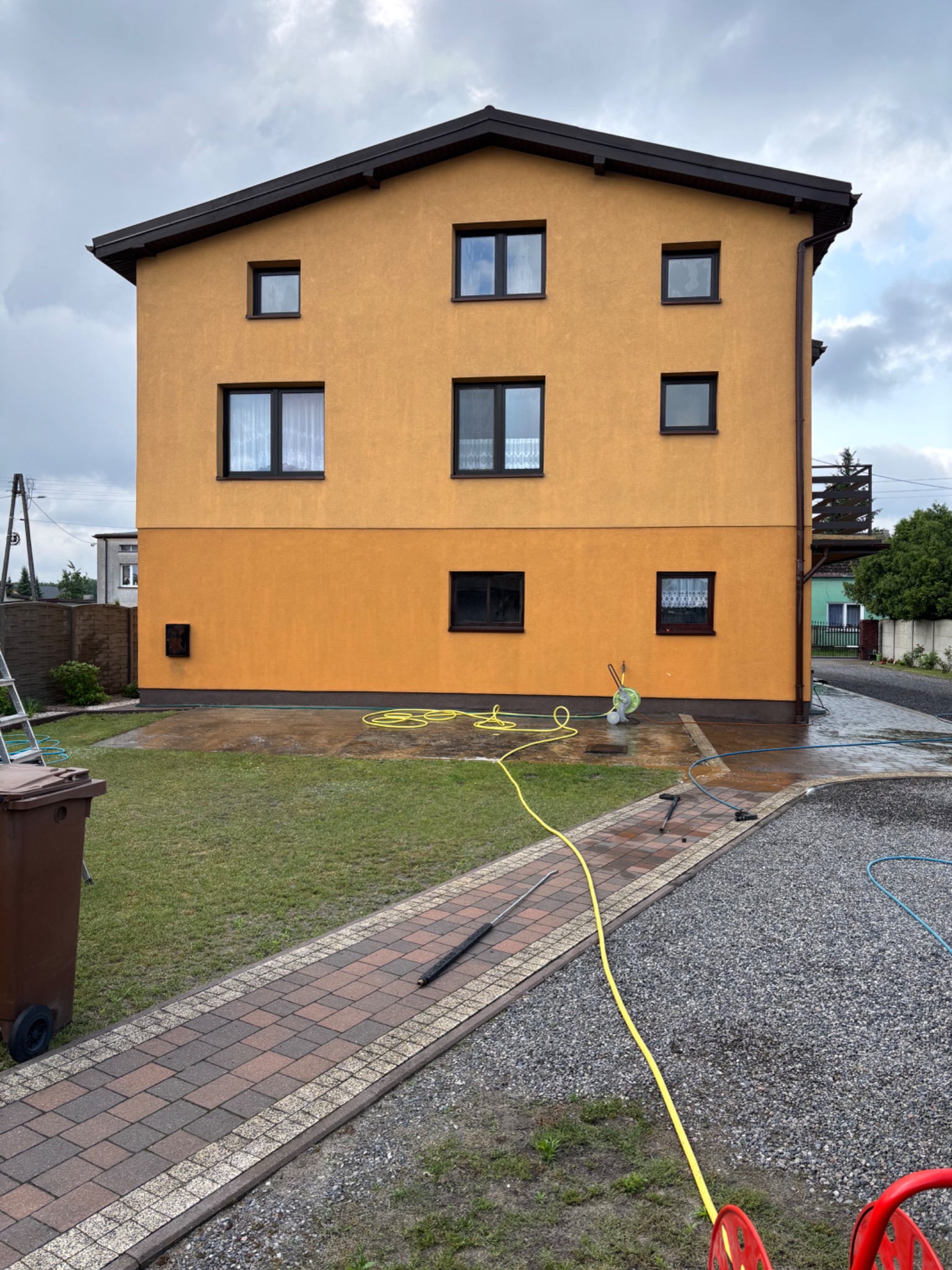 Dwupiętrowy dom z żółtą elewacją po myciu, z widocznym wężem i mokrym terenem wokół. Ślady wody na elewacji i chodniku.