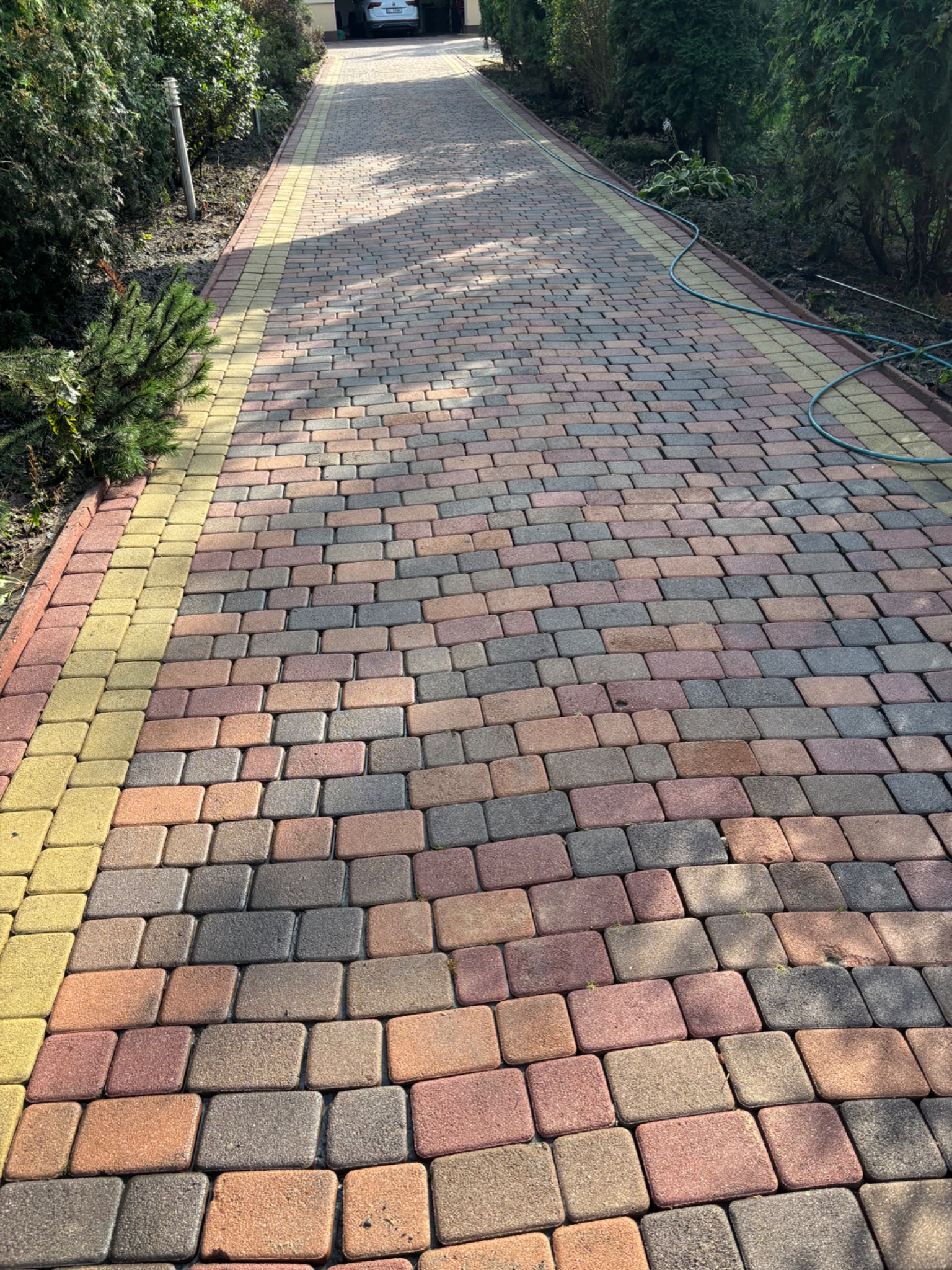 Kolorowa kostka brukowa na podjeździe, z żółtym obramowaniem, otoczona zielenią. W tle widoczny zaparkowany samochód i brama garażowa.