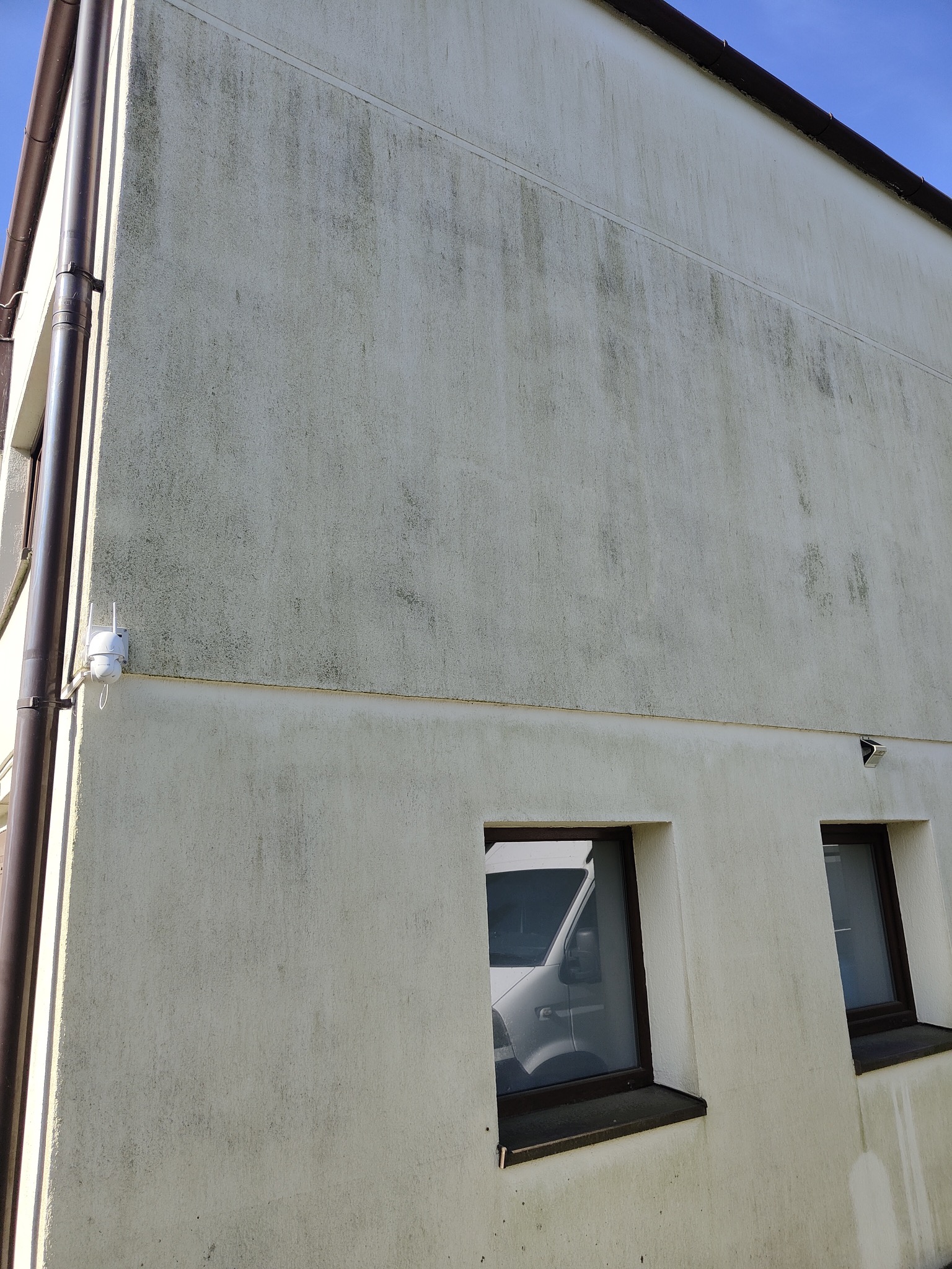 Brudna, jasna elewacja budynku z widocznymi zaciekami i zabrudzeniami, brązowe rynny, okna z odbijającym się samochodem. Ujęcie z dołu, niebieskie niebo.