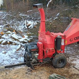 M&K usługi ziemne - Czerwona rębarka Cedrus na tle ściętych gałęzi i pni drzew pokrytych śniegiem, obok widoczna osoba w odblaskowej kurtce.