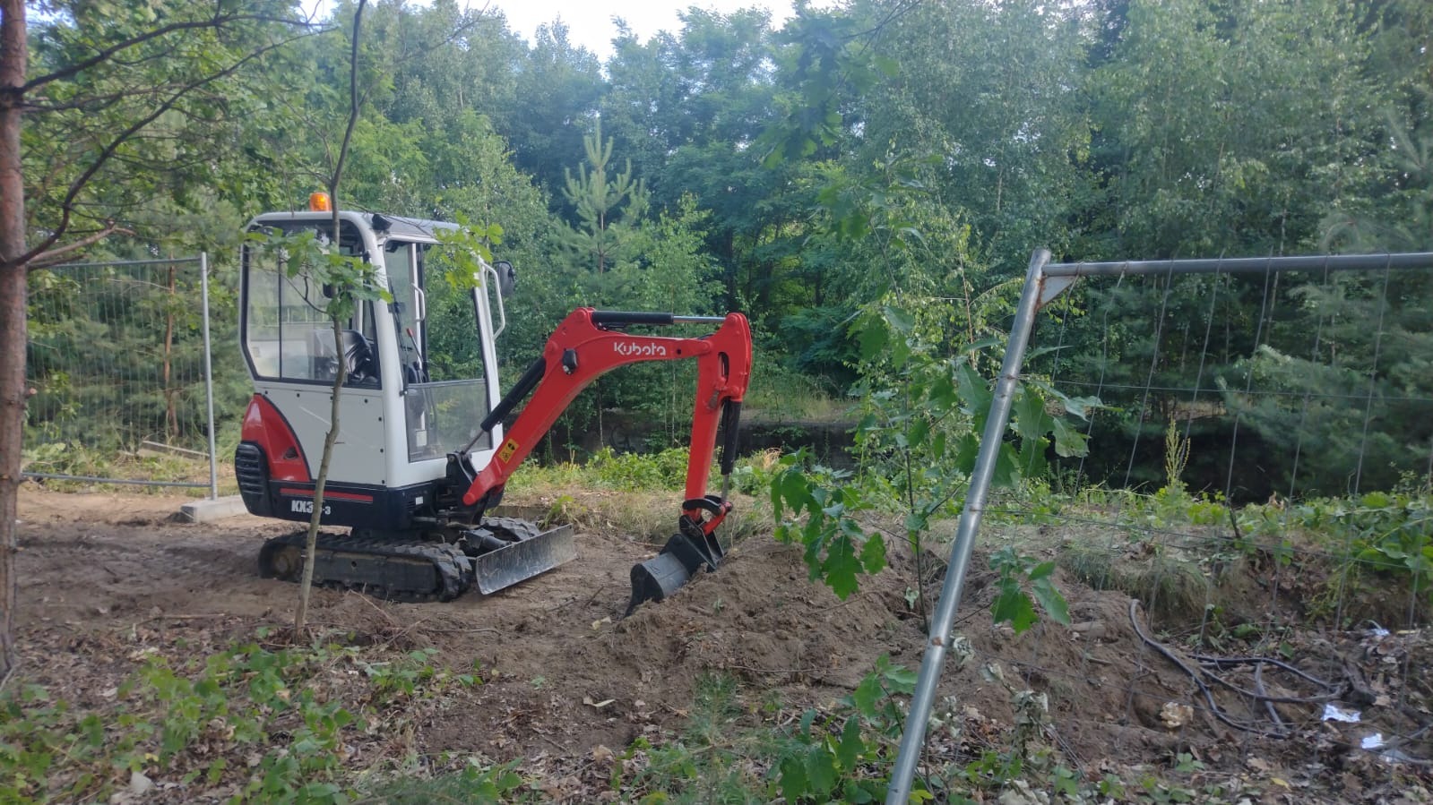 Mała koparka Kubota KX3-3 na placu budowy, widoczna świeżo wykopana ziemia i tymczasowe ogrodzenie z siatki, w tle zieleń drzew.