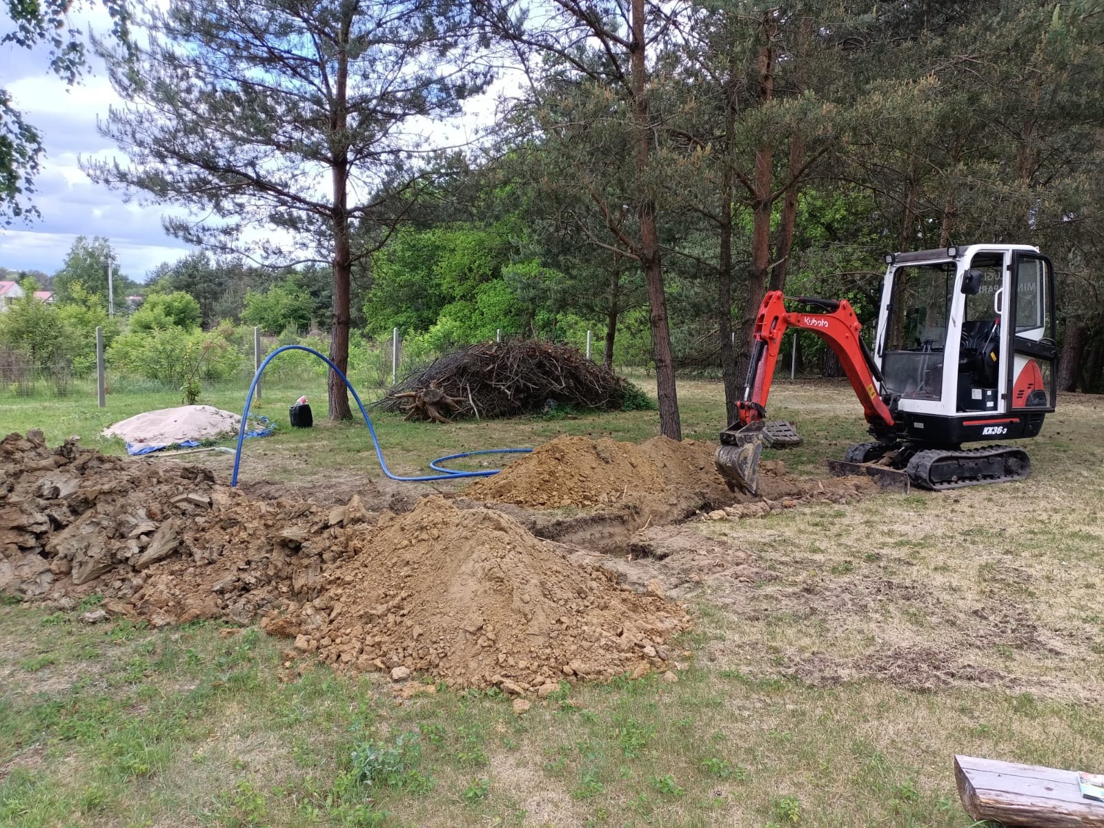 Mała koparka Kubota KX36-3 w trakcie wykonywania wykopu w ziemi, widoczny stos ziemi oraz niebieski wąż, w tle drzewa iglaste i sterta gałęzi.