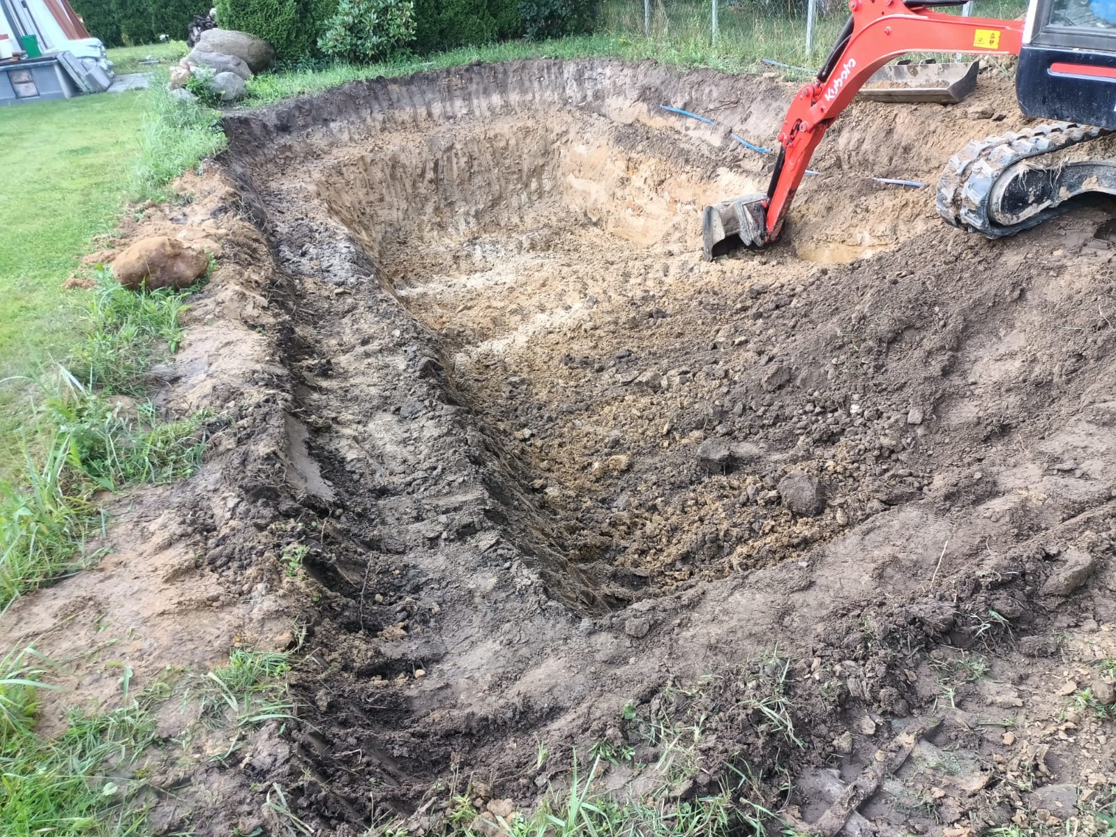 Czerwona minikoparka Kubota na placu budowy, podczas wykonywania głębokiego wykopu w ziemi, widoczne ślady gąsienic i ułożone rury.