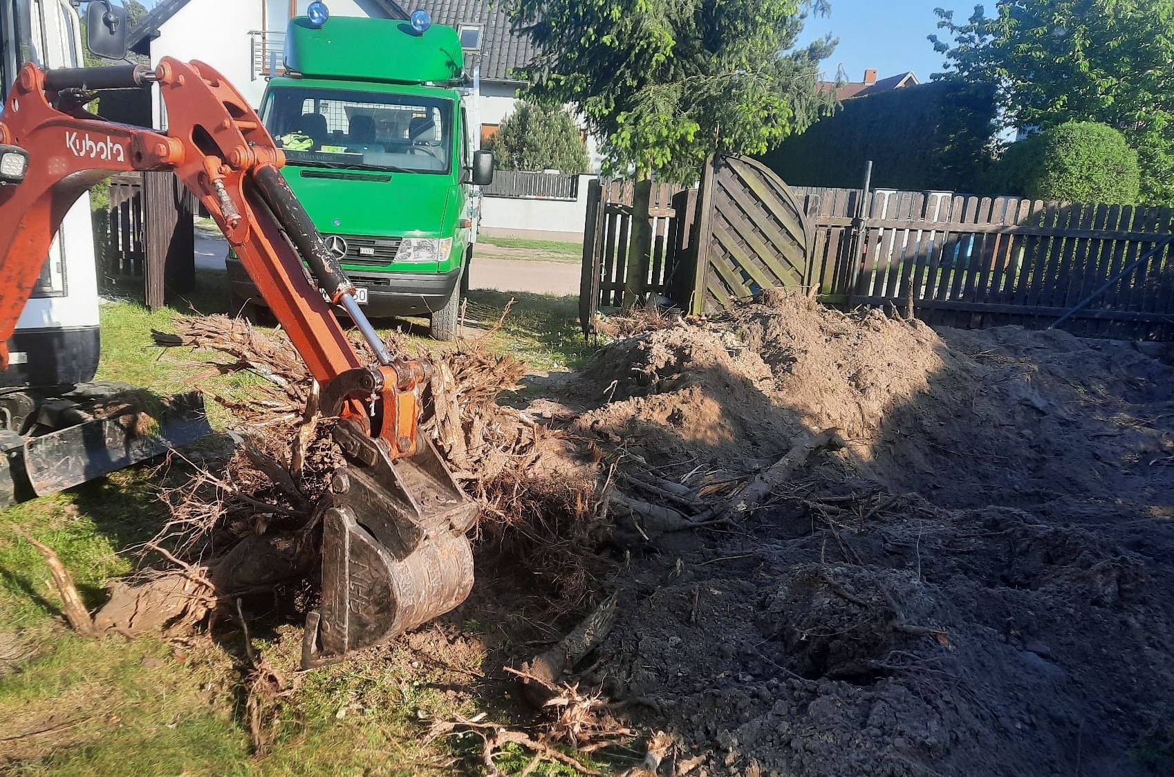 Pomarańczowa minikoparka Kubota z otwartą łyżką nabiera karpę po wyciętym drzewie, obok zielony samochód dostawczy Mercedes i drewniane ogrodzenie.