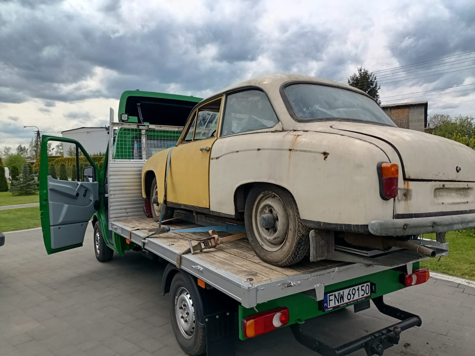 Uszkodzony, dwukolorowy (żółto-biały) samochód osobowy, zabezpieczony pasami transportowymi, przewożony na platformie lawety o zielonej kabinie, na tle pochmurnego nieba.