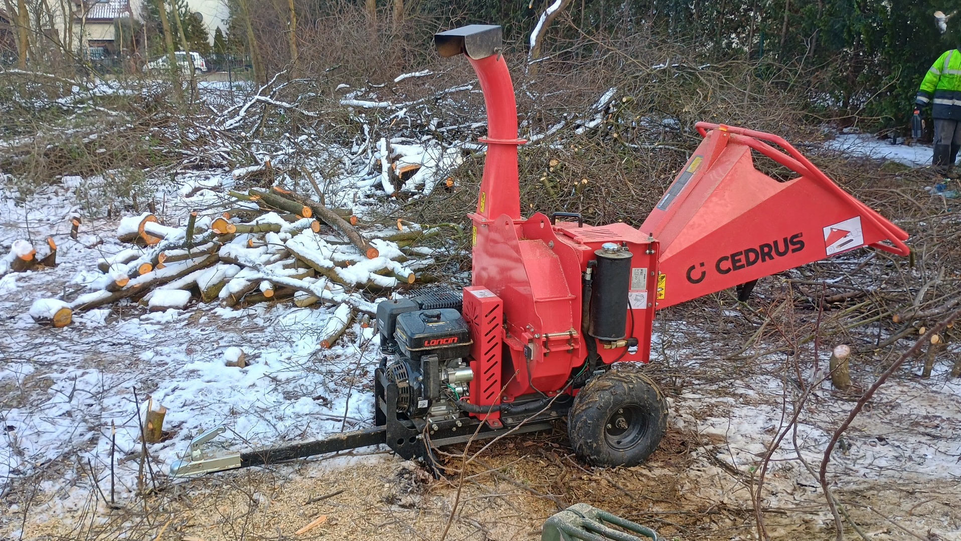 Czerwona rębarka Cedrus na tle ściętych gałęzi i pni drzew pokrytych śniegiem, obok widoczna osoba w odblaskowej kurtce.