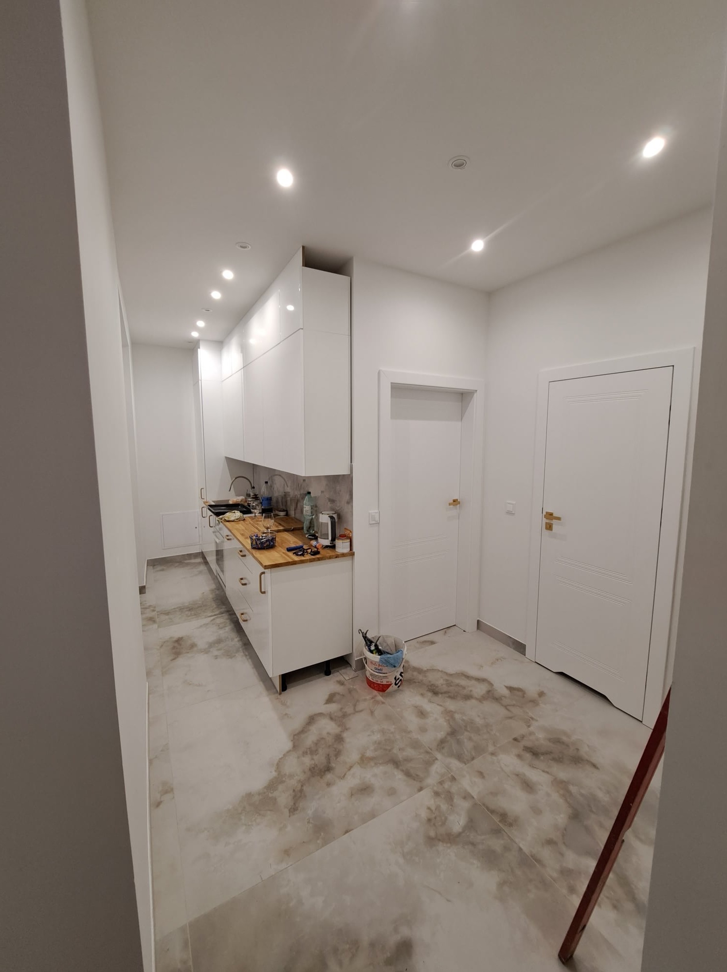 Jasne wnętrze kuchni w trakcie prac wykończeniowych, widoczne białe szafki, drewniany blat, marmurowe płytki podłogowe z widocznymi zabrudzeniami i ślady po farbie, białe drzwi i drabina oparta...