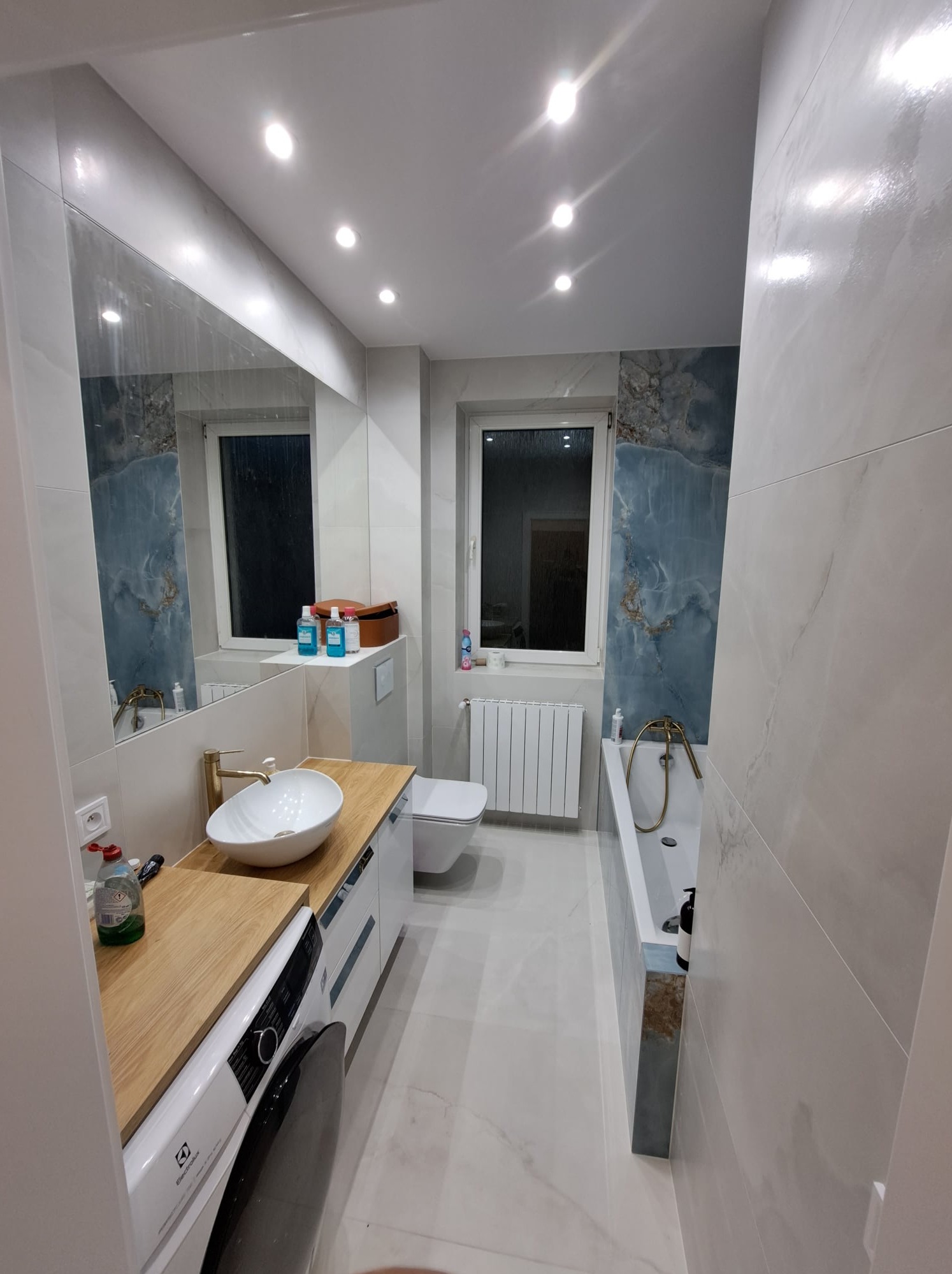 Nowoczesna łazienka z umywalką nablatową, białą armaturą, płytkami imitującymi marmur w odcieniach bieli i błękitu oraz drewnianym blatem, z pralką w zabudowie i oświetleniem punktowym.