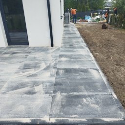 4you - Nowoczesna ścieżka z szarej kostki brukowej ułożona wzdłuż białego budynku. W tle widoczny pracownik podczas układania kostki i przygotowania terenu.