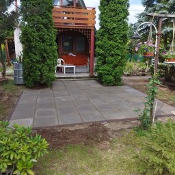 4you - Szare płyty chodnikowe tworzą patio przed domkiem z balkonem, otoczone zielenią. Widoczne tuje, kwiaty w wiszących donicach i fragment trawnika. Obrzeża patio wykończone brązową listwą.