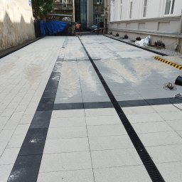 4you - Nowa nawierzchnia z jasnoszarej kostki brukowej z ciemnymi pasami i odpływem liniowym, teren budowy w tle, częściowo ukończona realizacja.