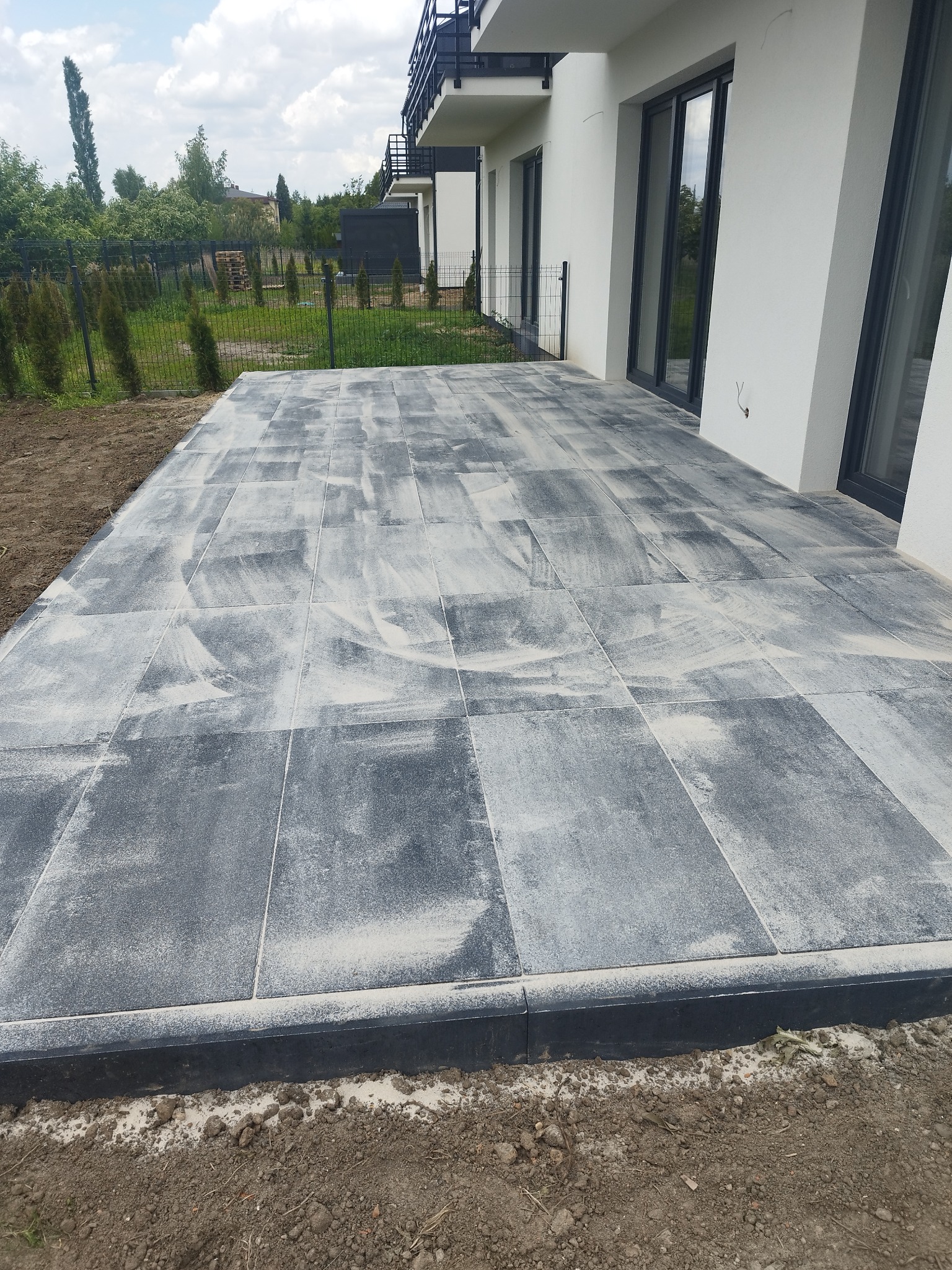 Nowoczesny taras z szarej kostki brukowej przy budynku w Łodzi. Widoczne świeżo ułożone płyty i obrzeża. Minimalistyczny design, jasna elewacja budynku.