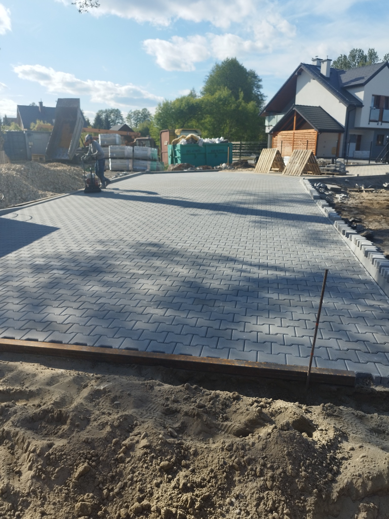Szara kostka brukowa ułożona na podjeździe domu jednorodzinnego. W tle widoczny pracownik zagęszczający podłoże. Częściowo ułożone obrzeża.