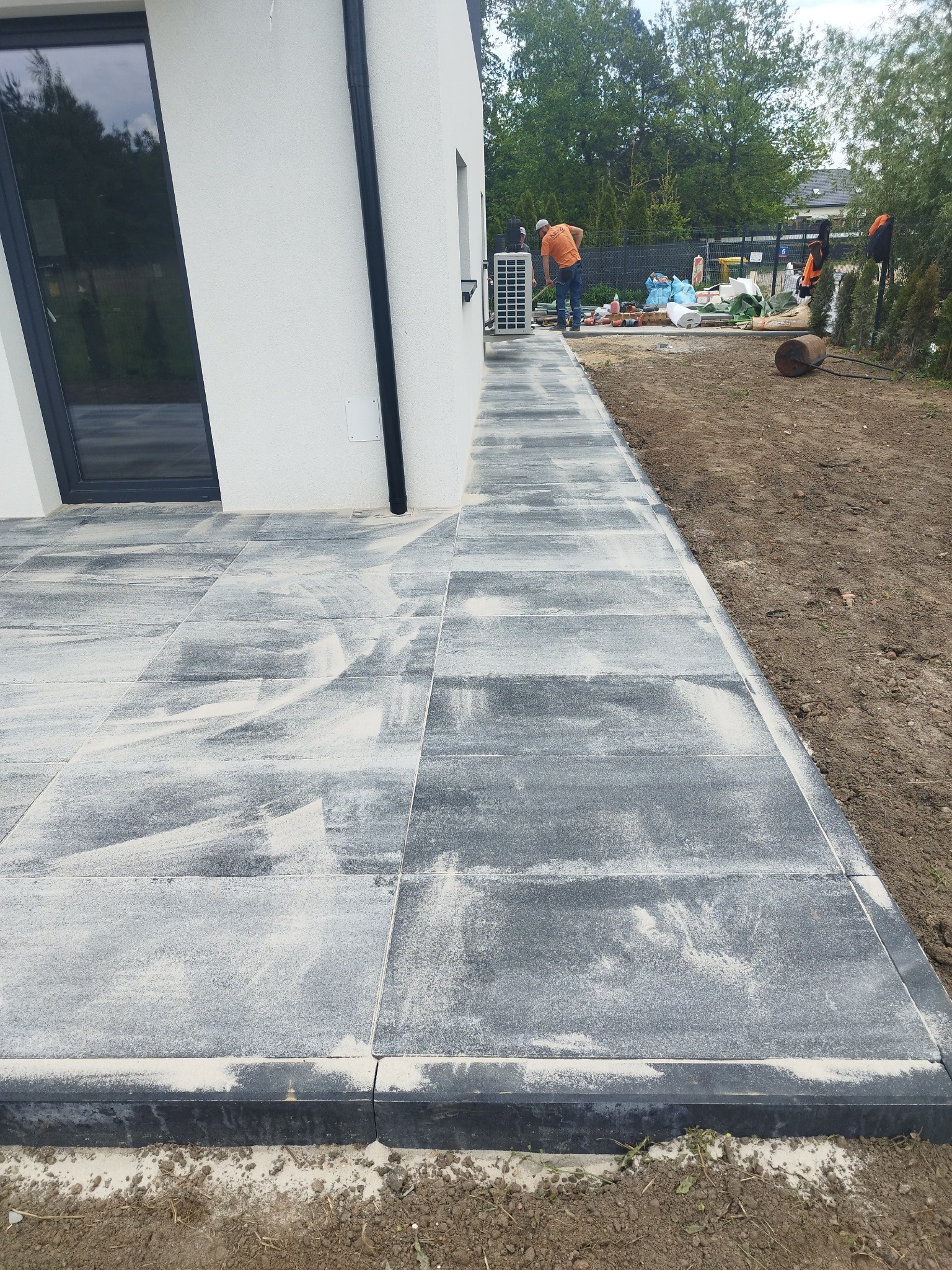 Nowoczesna ścieżka z szarej kostki brukowej ułożona wzdłuż białego budynku. W tle widoczny pracownik podczas układania kostki i przygotowania terenu.