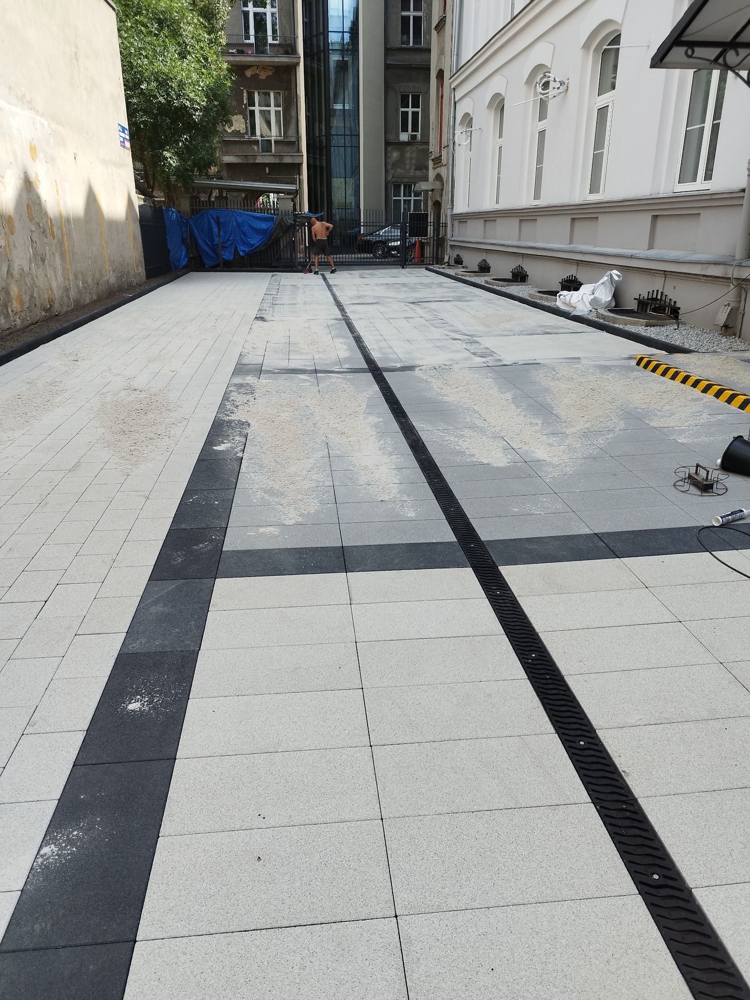 Nowa nawierzchnia z jasnoszarej kostki brukowej z ciemnymi pasami i odpływem liniowym, teren budowy w tle, częściowo ukończona realizacja.