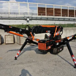 Czarno-pomarańczowy pająk-dźwig BG Lift M250 z wysuniętymi podporami, na tle betonowego muru i metalowej konstrukcji z drewnianymi panelami.