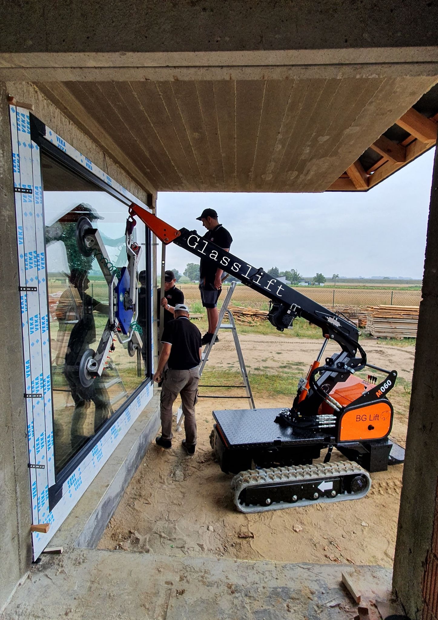 Montaż okna za pomocą mini dźwigu gąsienicowego Glasslift BG Lift M060, trzech pracowników i drabiny, widok z zewnątrz budynku w trakcie budowy.