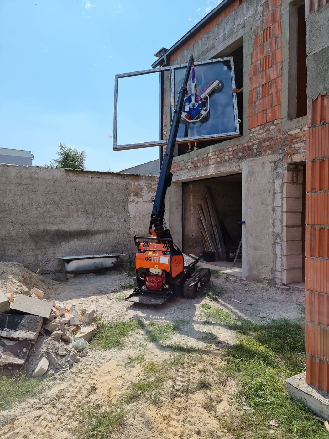 Mały, pomarańczowy dźwig gąsienicowy marki BG Lift podnosi okno z ramą na budowie domu z czerwonej cegły. W tle widoczny gruz, stara wanna i fragment szarego muru.