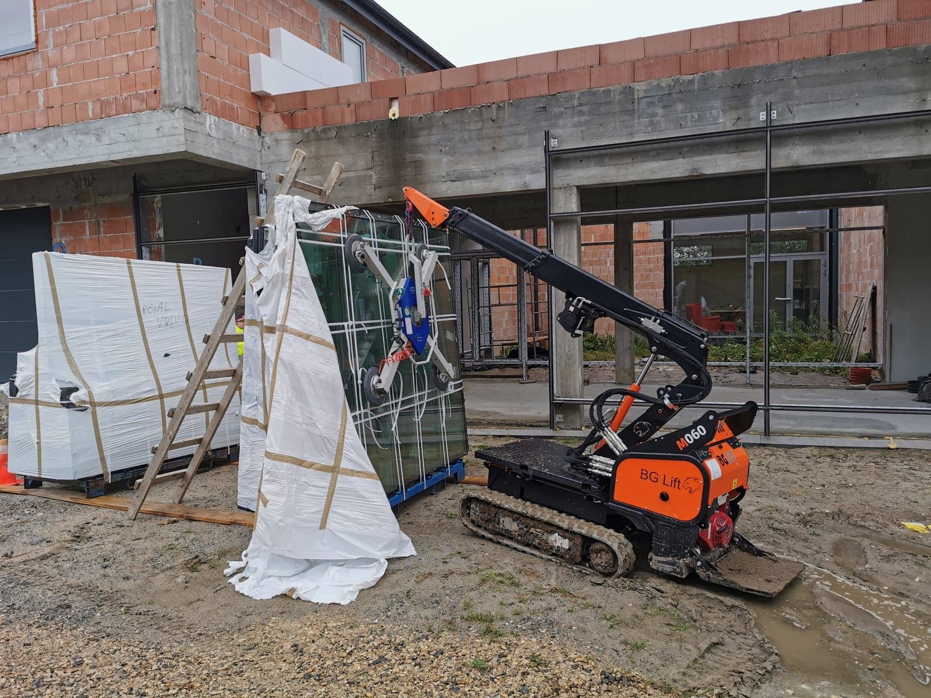 Mały dźwig gąsienicowy BG Lift M060 transportuje szybę na budowie domu w stanie surowym, obok stoją zapakowane szyby i drabina