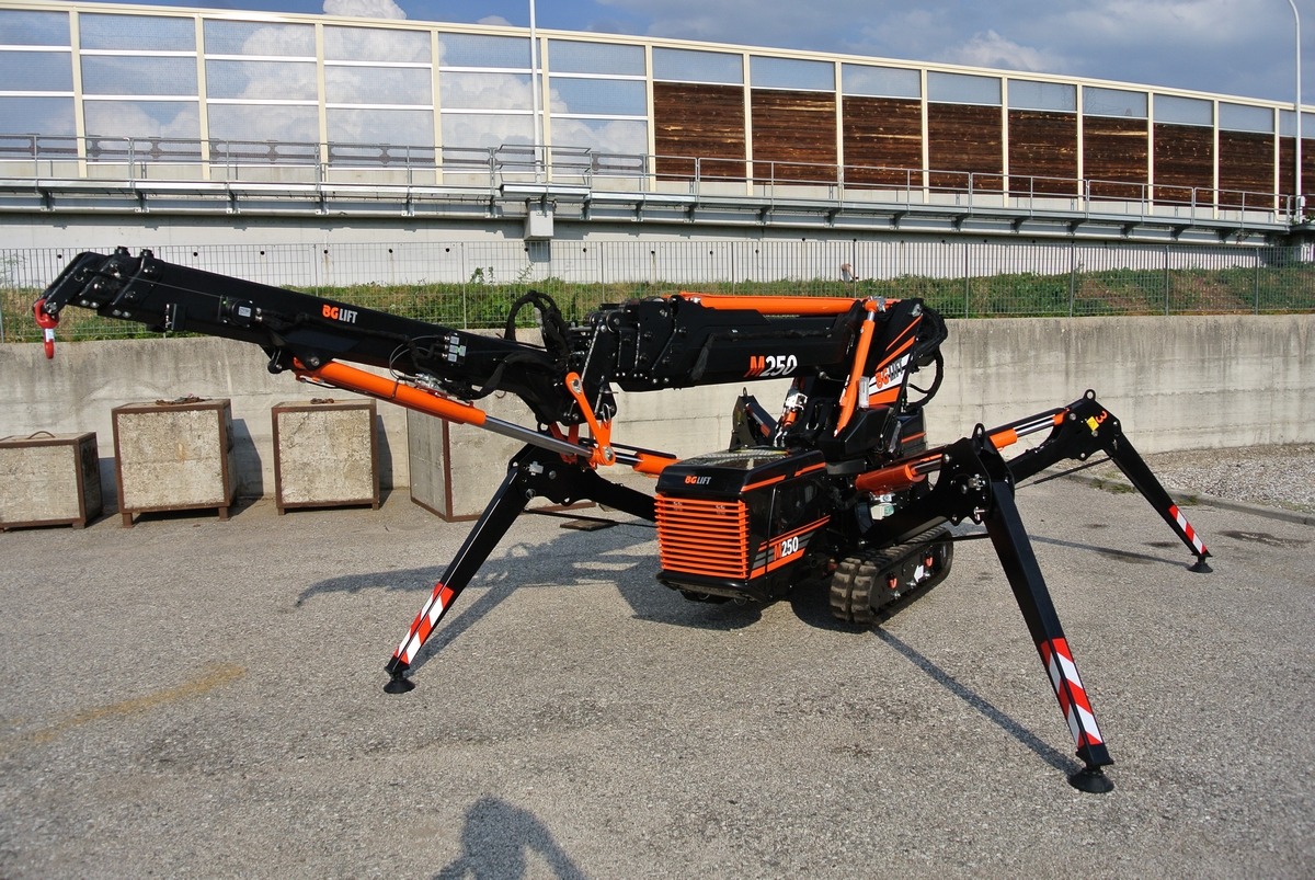 Czarno-pomarańczowy pająk-dźwig BG Lift M250 z wysuniętymi podporami, na tle betonowego muru i metalowej konstrukcji z drewnianymi panelami.