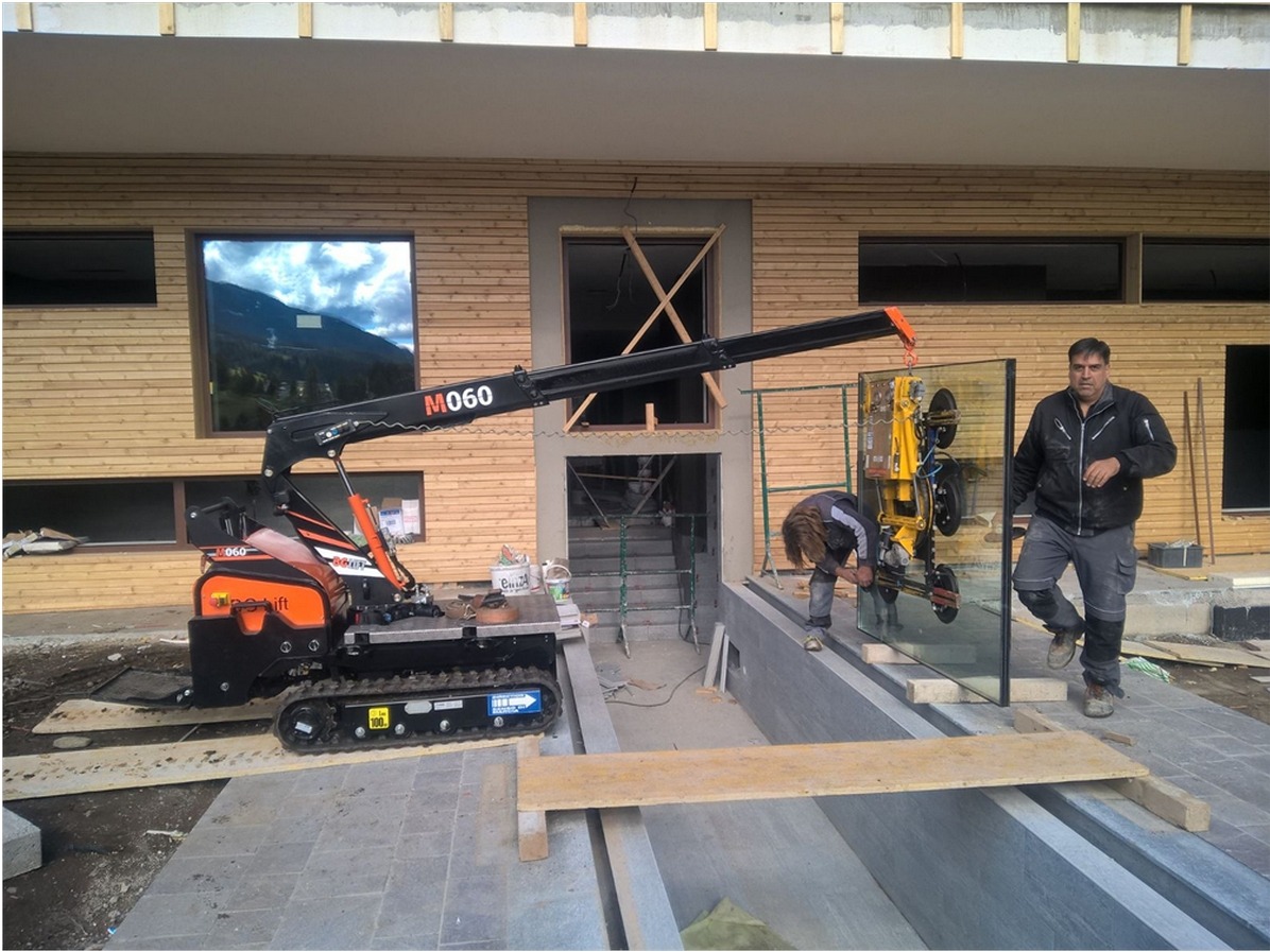 Montaż okna z użyciem mini-dźwigu gąsienicowego M060 na tle drewnianej elewacji budynku. Dwóch pracowników przy pracy, jeden stabilizuje szybę, drugi nadzoruje proces.