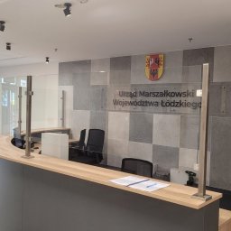 PKZ nad Łódką Sp. z o.o. - Nowoczesna recepcja w biurze Urzędu Marszałkowskiego Województwa Łódzkiego z przeszkloną osłoną, wykończona płytkami w odcieniach szarości i drewnianymi elementami dekoracyjnymi.