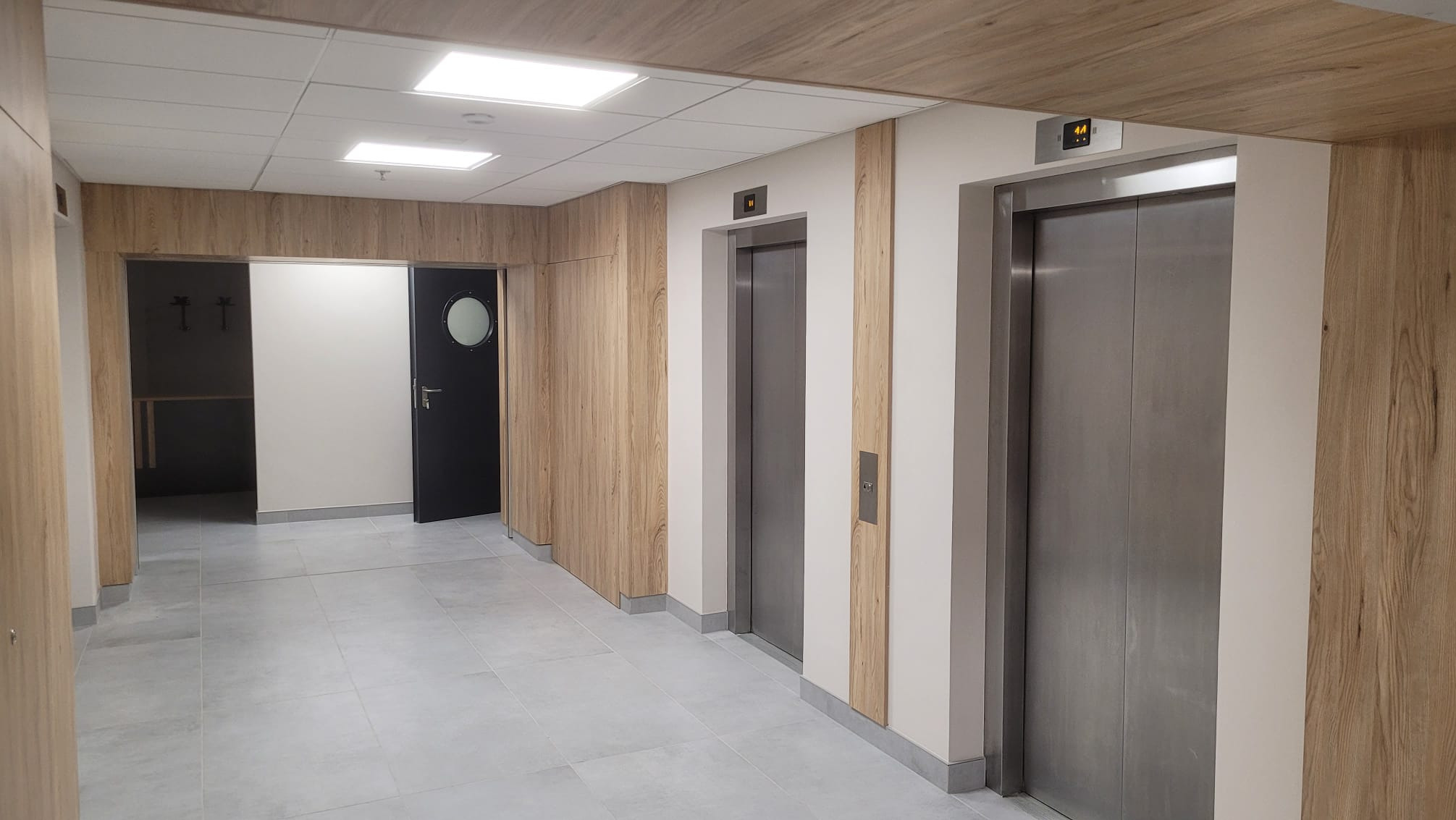 Wnętrze korytarza z dwoma windami, wykończone panelami drewnopodobnymi i szarą płytką podłogową. Jedna winda pokazuje numer piętra '44'.