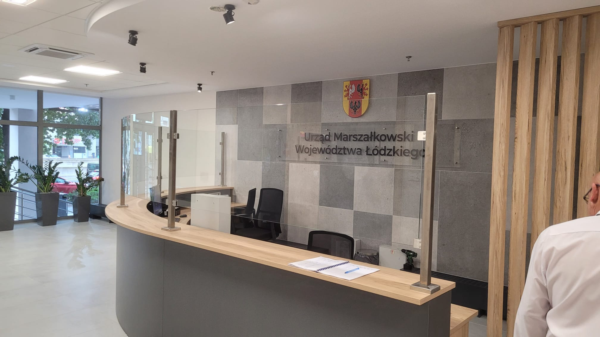 Nowoczesna recepcja w biurze Urzędu Marszałkowskiego Województwa Łódzkiego z przeszkloną osłoną, wykończona płytkami w odcieniach szarości i drewnianymi elementami dekoracyjnymi.