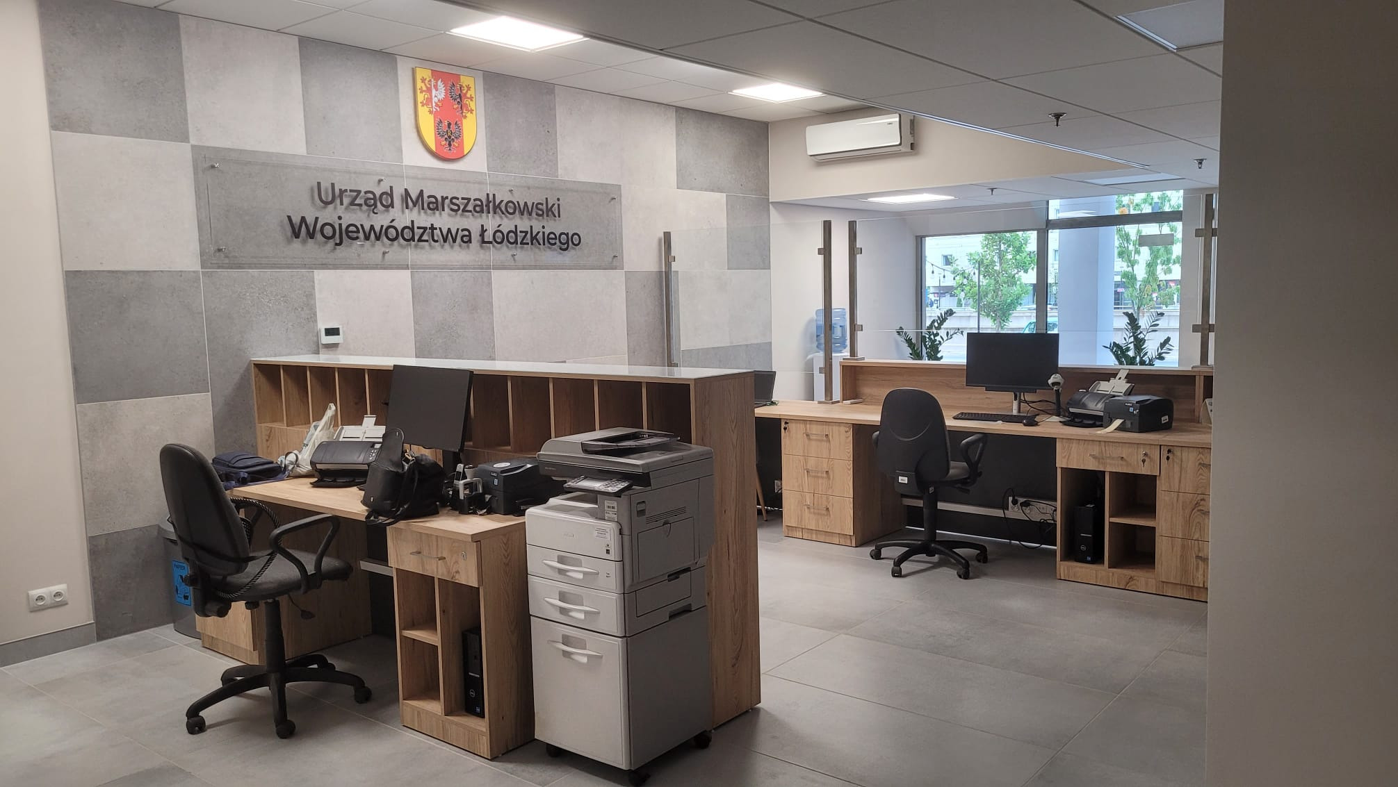 Nowoczesne wnętrze biurowe z recepcją, ściana wykończona płytkami imitującymi beton, logo Urzędu Marszałkowskiego Województwa Łódzkiego, drewniane biurka i krzesła biurowe, widok na zieleń za oknem.