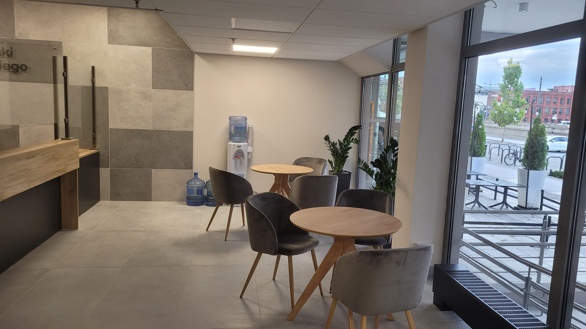 Nowoczesne wnętrze biurowe z recepcją z drewnianym blatem, strefą relaksu z okrągłymi stolikami i szarymi krzesłami, dystrybutorem wody i widokiem na ulicę.