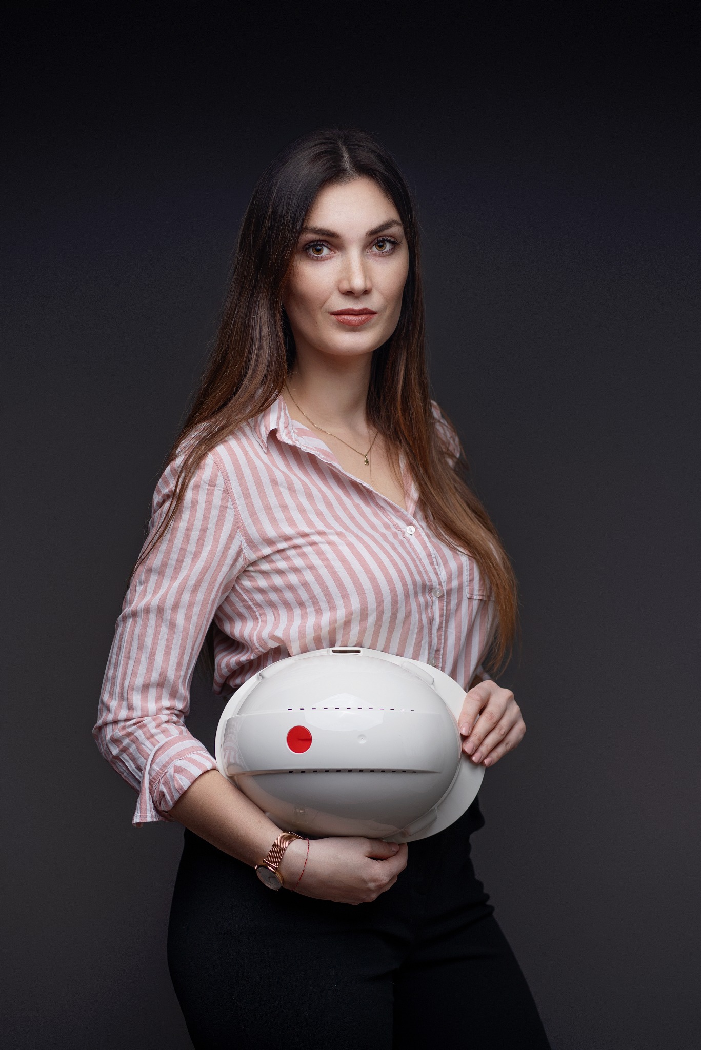 Kobieta w różowo-białej koszuli trzyma biały kask ochronny z czerwoną naklejką na ciemnoszarym tle.