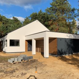 BUDMAN SPÓŁKA Z OGRANICZONĄ ODPOWIEDZIALNOŚCIĄ - SPÓŁKA KOMANDYTOWA - Nowoczesny, parterowy dom z płaskim dachem i garażem w trakcie wykańczania terenu, z widocznymi kostkami brukowymi na palecie i małą łopatką w piasku.