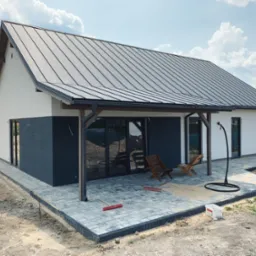 Nowoczesny dom z szarą blachą na dachu, częściowo wykończony, z drewnianymi krzesłami i brukowanym tarasem w trakcie budowy.