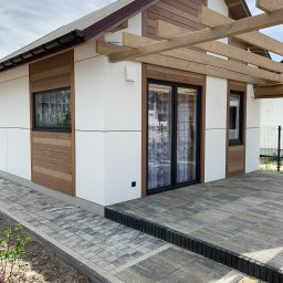 BUDMAN SPÓŁKA Z OGRANICZONĄ ODPOWIEDZIALNOŚCIĄ - SPÓŁKA KOMANDYTOWA - Nowoczesny, parterowy dom z białymi panelami i drewnianą elewacją, z dużymi przesuwnymi drzwiami tarasowymi i drewnianą konstrukcją pergoli nad tarasem wyłożonym kostką brukową.