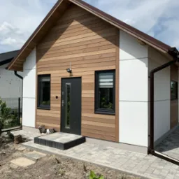 Nowoczesny dom z drewnianą elewacją i białymi panelami, ciemne okna i drzwi, kostka brukowa wokół, widoczne elementy instalacji odwadniającej.