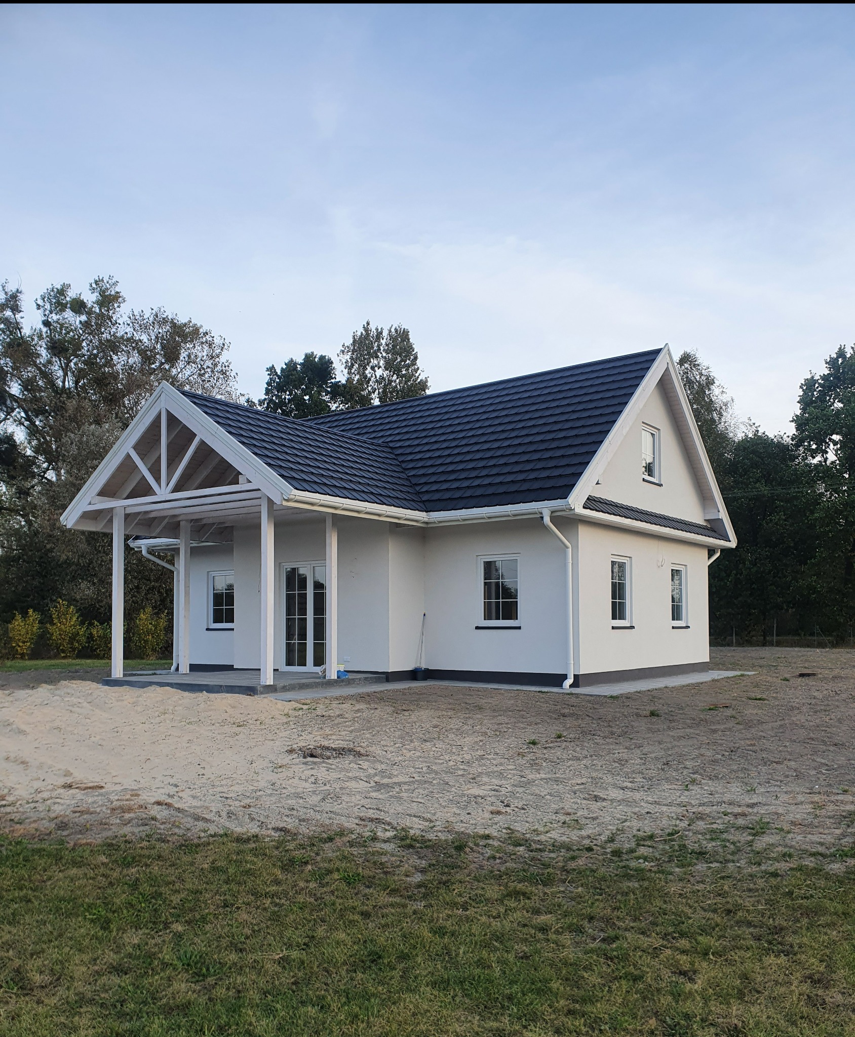 Nowoczesny, parterowy dom z białymi ścianami i antracytowym dachem, zadaszony taras z drewnianą konstrukcją, widok z zewnątrz na jeszcze niezagospodarowany teren.