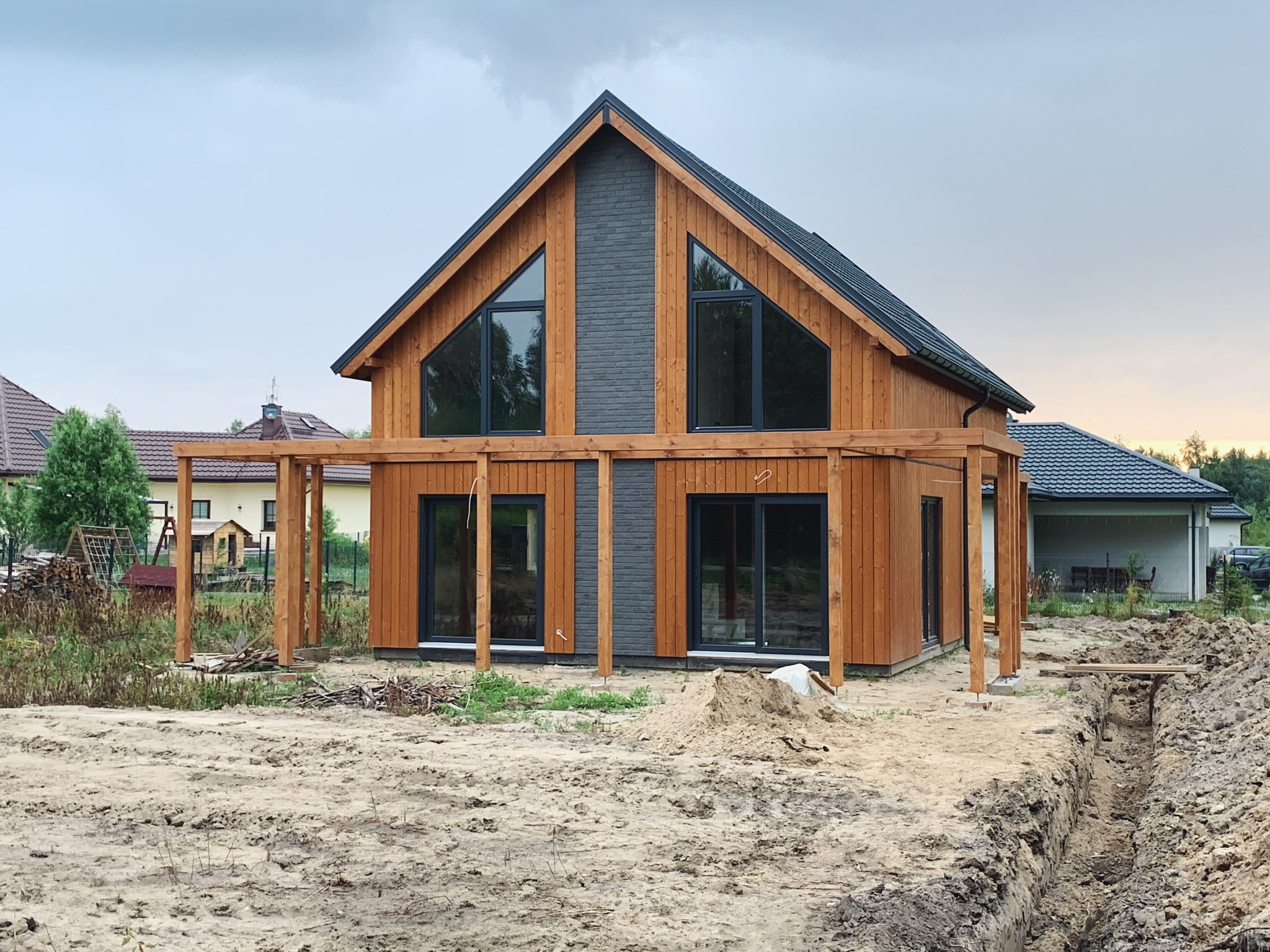 Nowoczesny dom z drewna z elementami szarej cegły na szczycie, z dużymi oknami i konstrukcją drewnianą pergoli w trakcie budowy, widoczny wykop w ziemi.