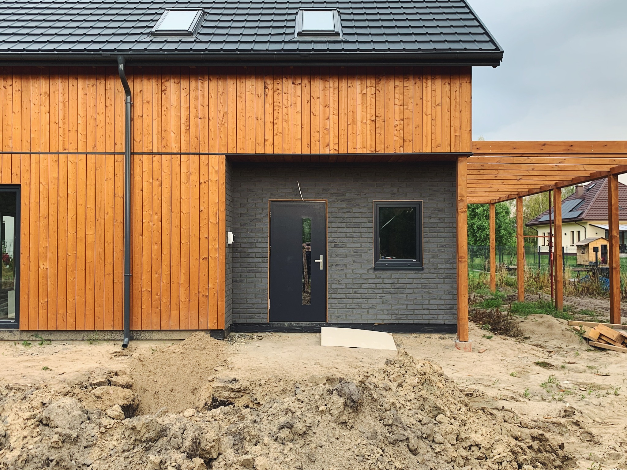 Nowoczesny dom szkieletowy z elewacją z jasnego drewna, grafitową cegłą, czarną stolarką okienną i pergolą, w trakcie prac wykończeniowych wokół budynku.