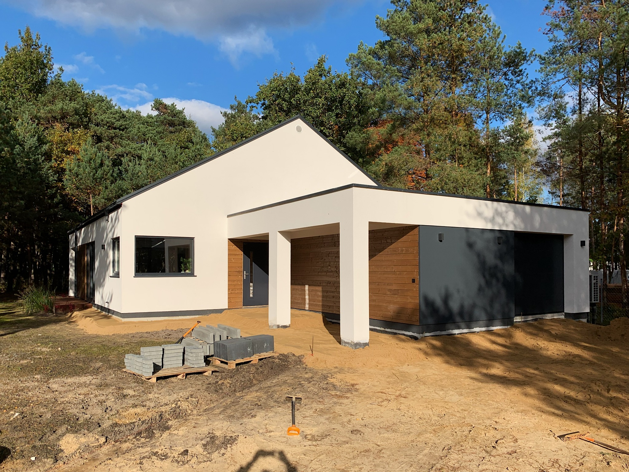 Nowoczesny, parterowy dom z płaskim dachem i garażem w trakcie wykańczania terenu, z widocznymi kostkami brukowymi na palecie i małą łopatką w piasku.