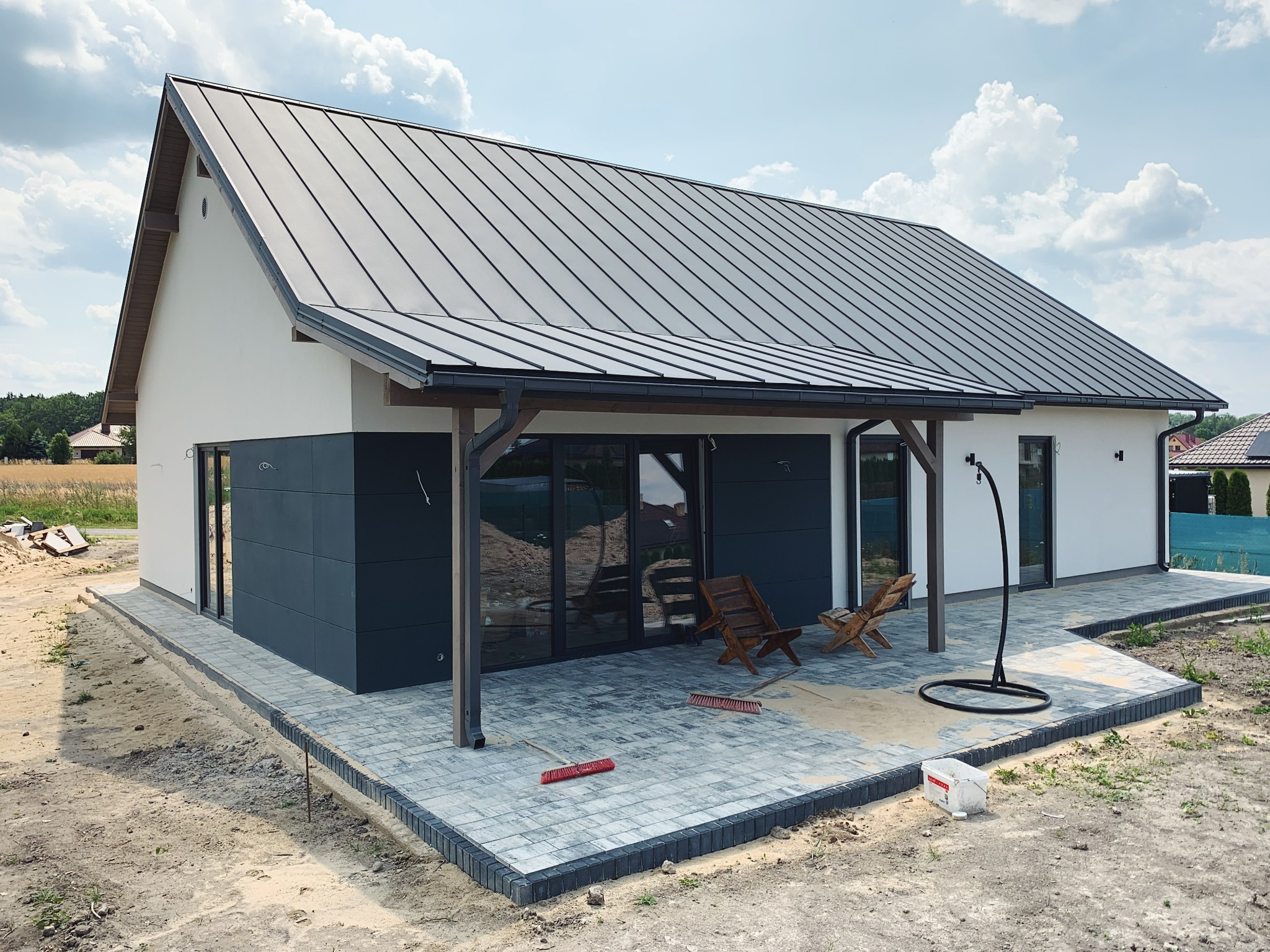 Nowoczesny dom z szarą blachą na dachu, częściowo wykończony, z drewnianymi krzesłami i brukowanym tarasem w trakcie budowy.