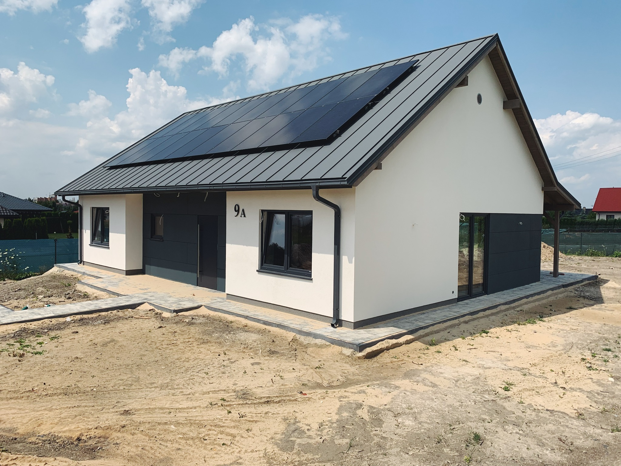 Nowoczesny, parterowy dom z panelami słonecznymi na szarym, metalowym dachu i jasną elewacją, numer 9A nad drzwiami wejściowymi, otoczony piaskiem i częściowo ułożoną kostką brukową.
