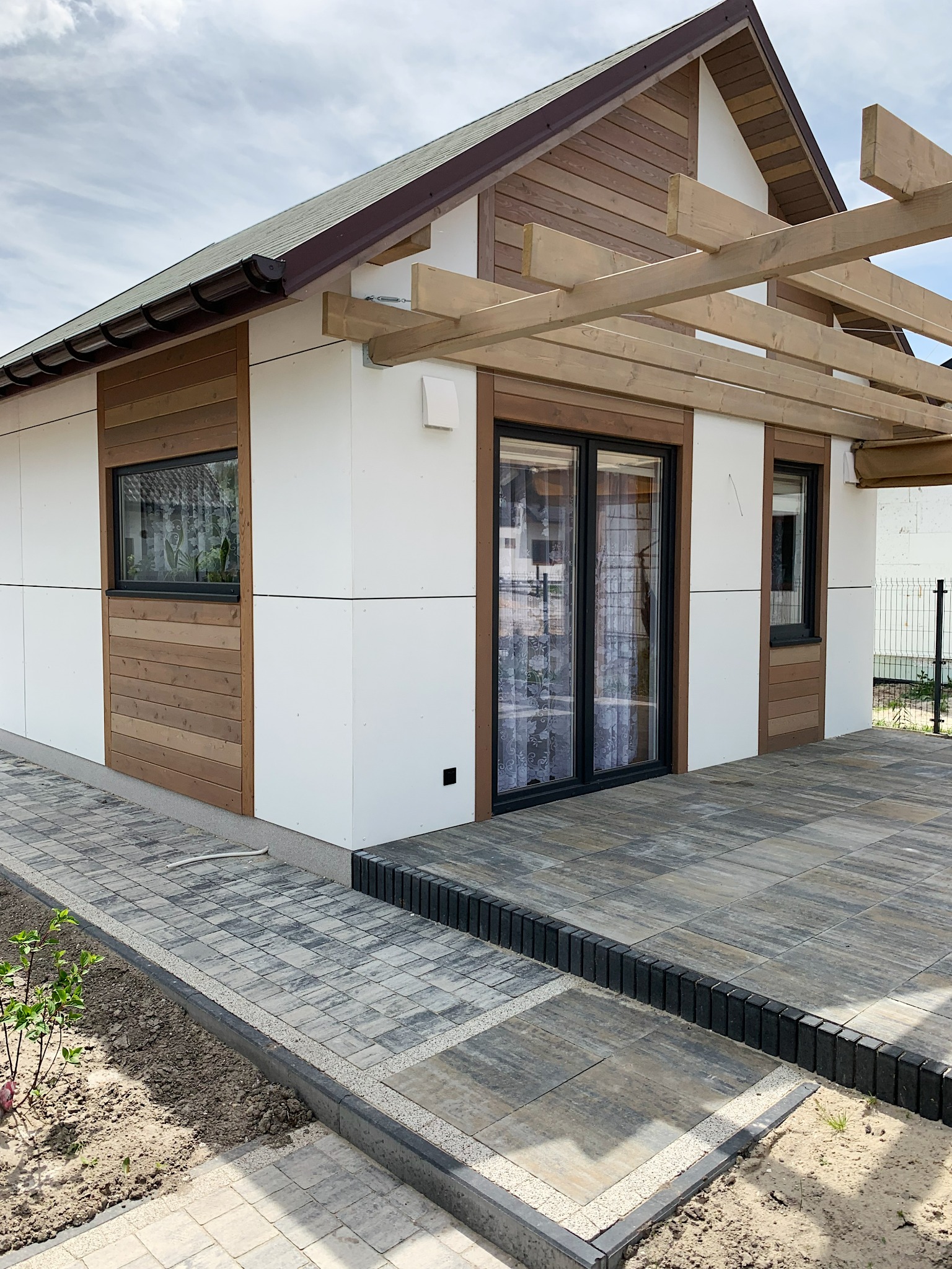 Nowoczesny, parterowy dom z białymi panelami i drewnianą elewacją, z dużymi przesuwnymi drzwiami tarasowymi i drewnianą konstrukcją pergoli nad tarasem wyłożonym kostką brukową.