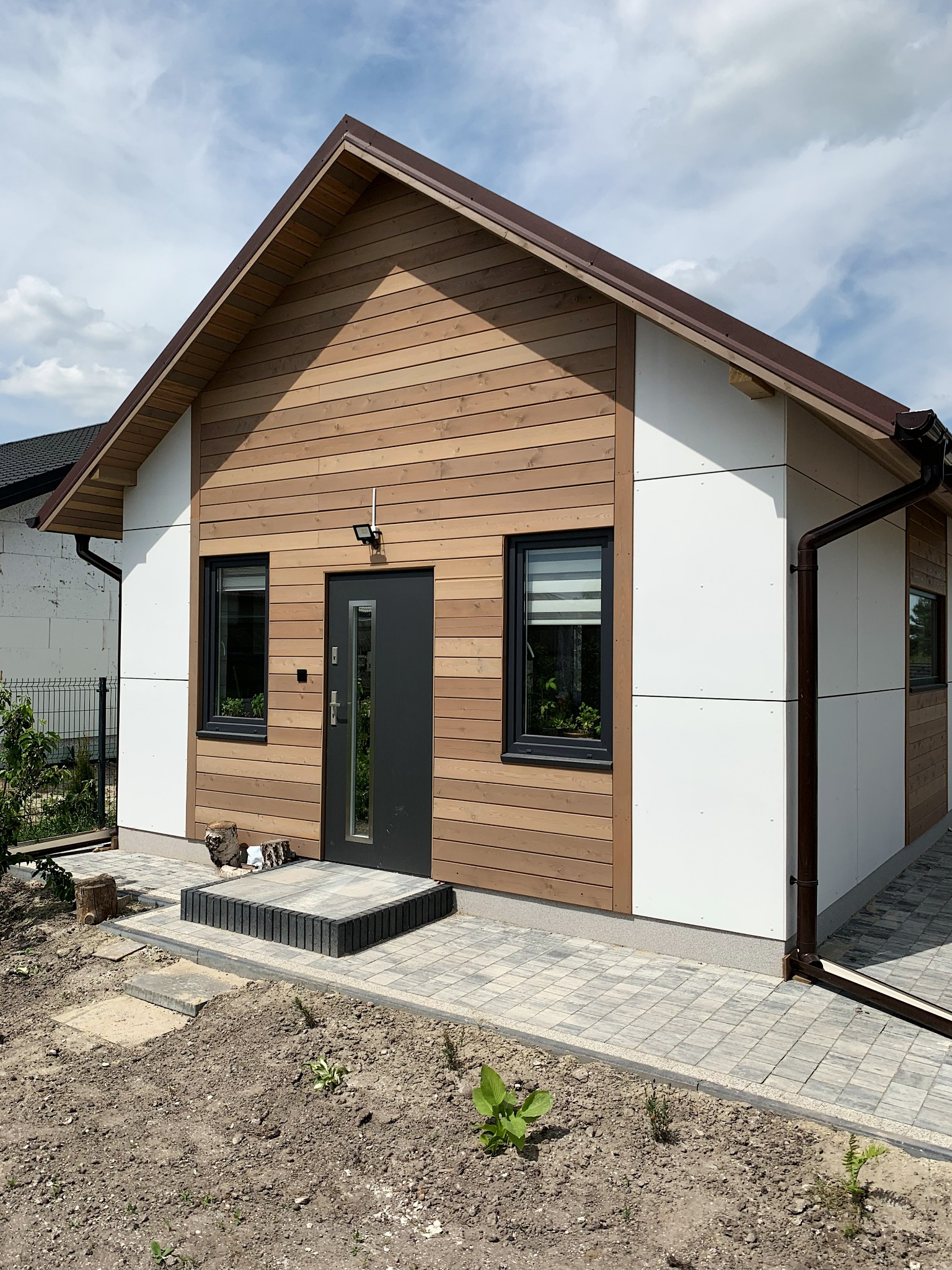 Nowoczesny dom z drewnianą elewacją i białymi panelami, ciemne okna i drzwi, kostka brukowa wokół, widoczne elementy instalacji odwadniającej.