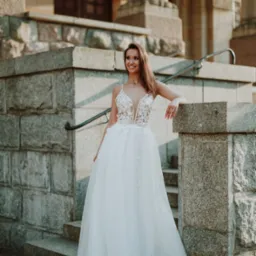 Uśmiechnięta kobieta w białej, eleganckiej sukni z koronkową górą, stojąca na kamiennych schodach przed budynkiem z kolumnami i łukami.