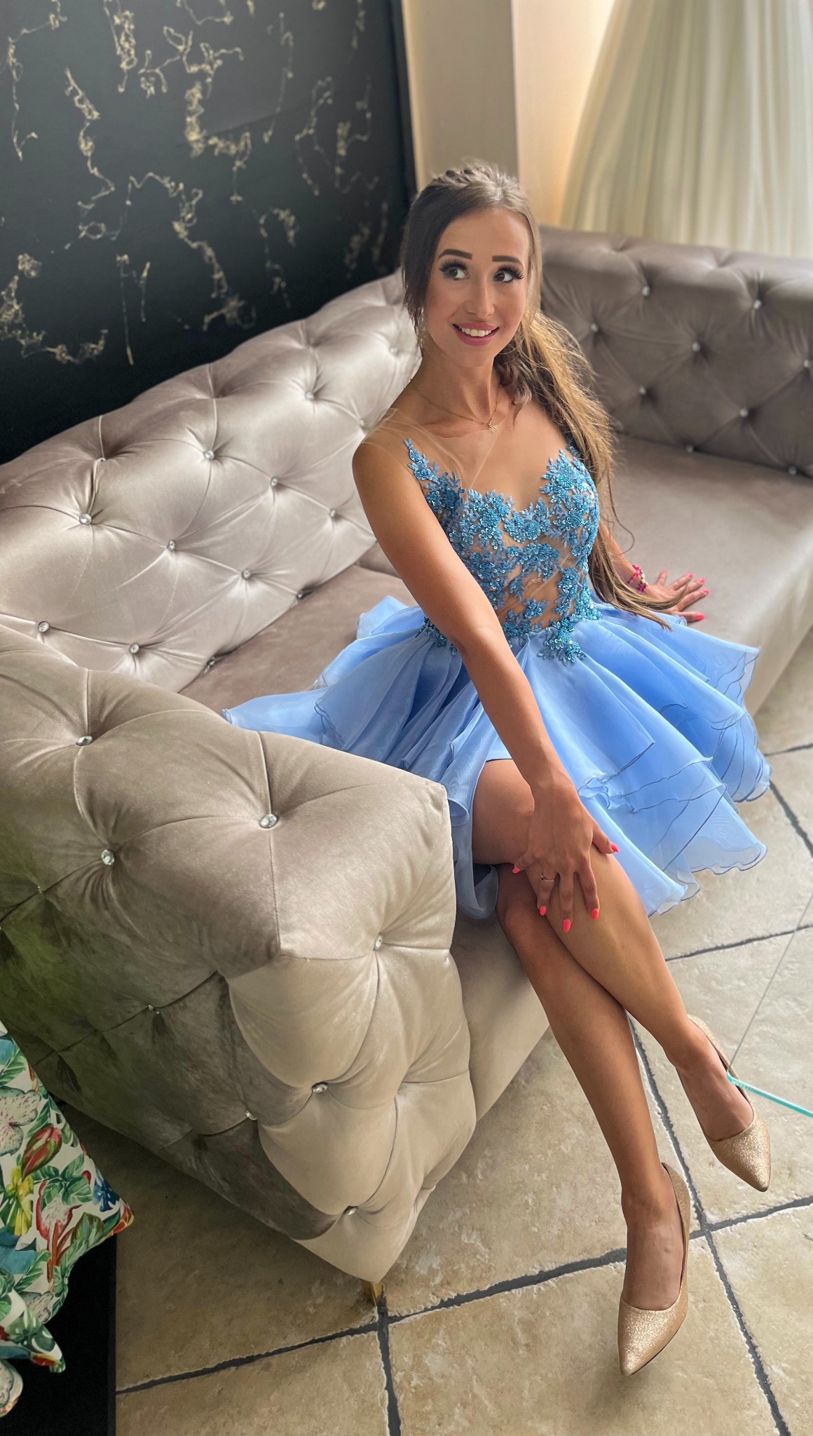 Uśmiechnięta kobieta siedzi na pikowanej sofie w błękitnej, warstwowej sukience z koronkową górą, prezentując strój i złote szpilki.