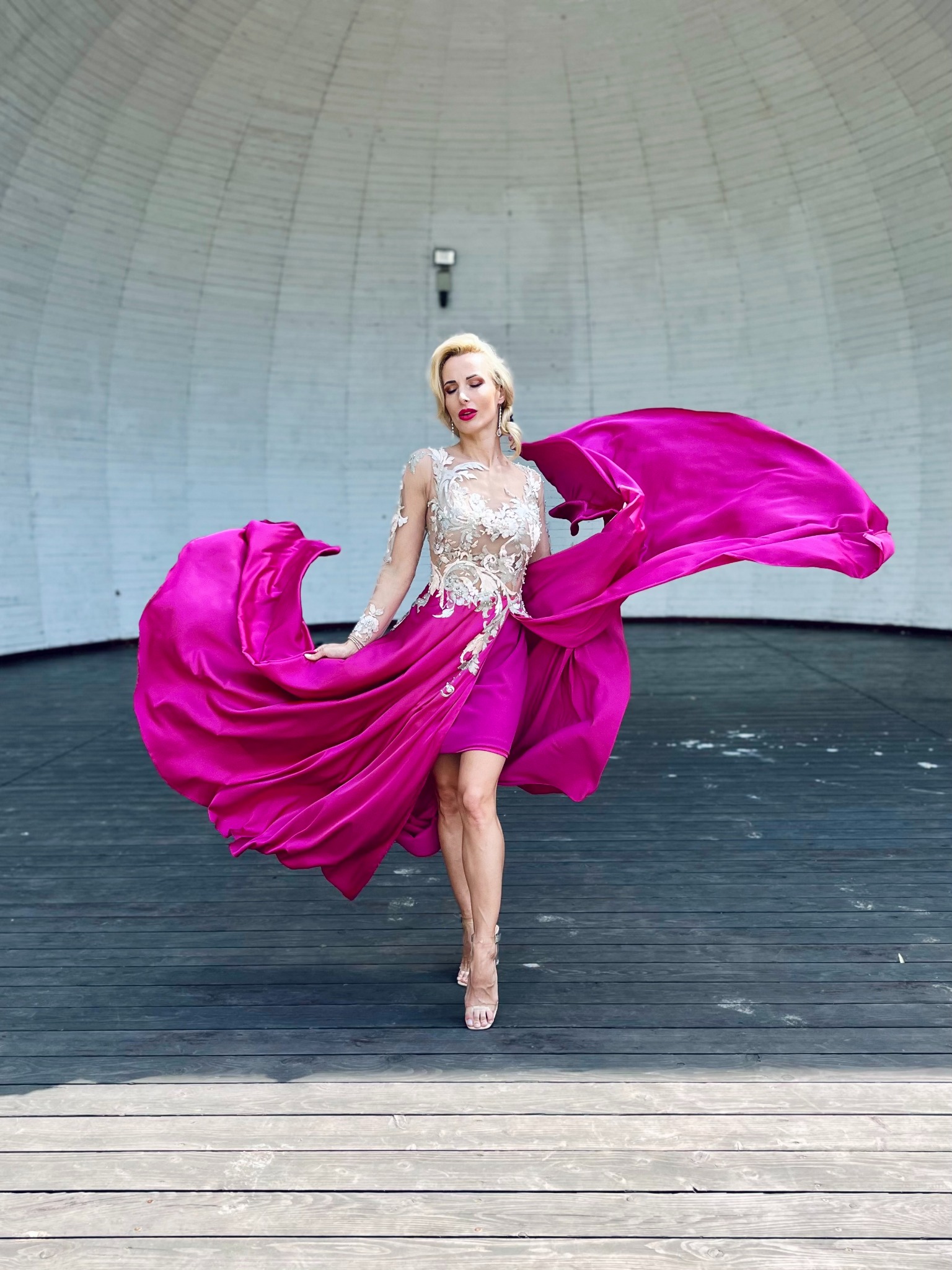 Blondwłosa kobieta w krótkiej sukience z koronkową górą i obszerną, fuksjową spódnicą, pozuje na drewnianej scenie pod zadaszeniem.