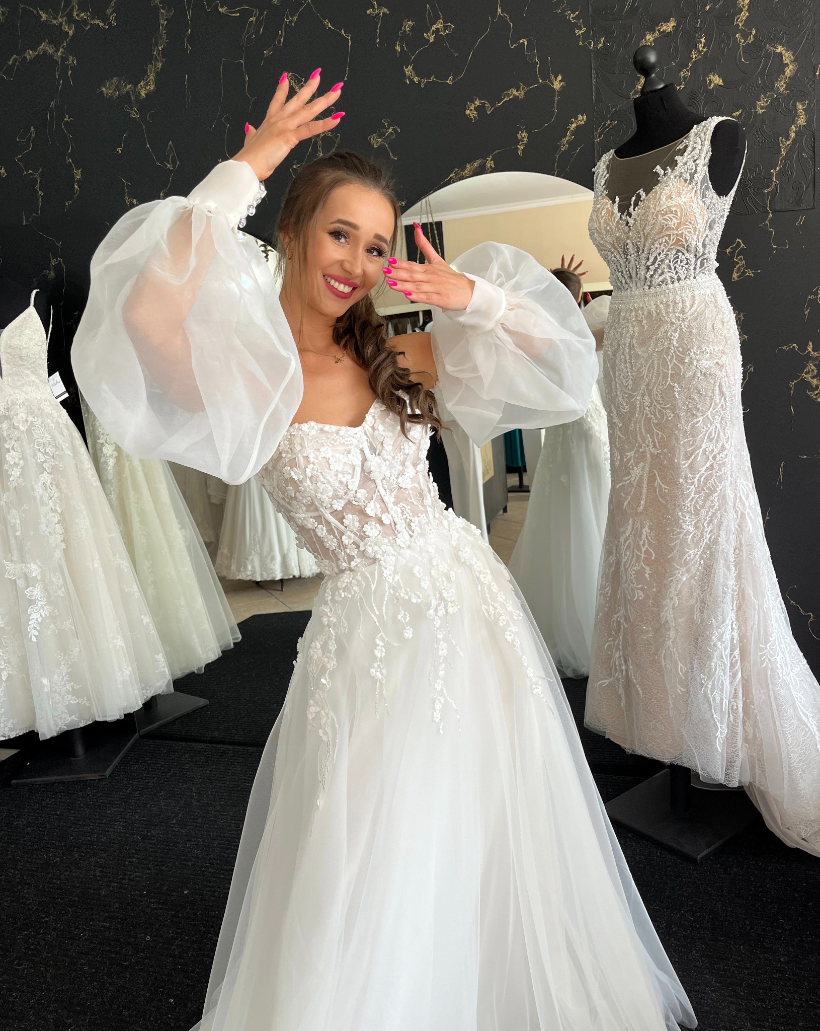 Uśmiechnięta kobieta w koronkowej sukni ślubnej z bufiastymi rękawami pozuje w salonie, na tle innych sukien i czarnej ściany ze złotymi zdobieniami.