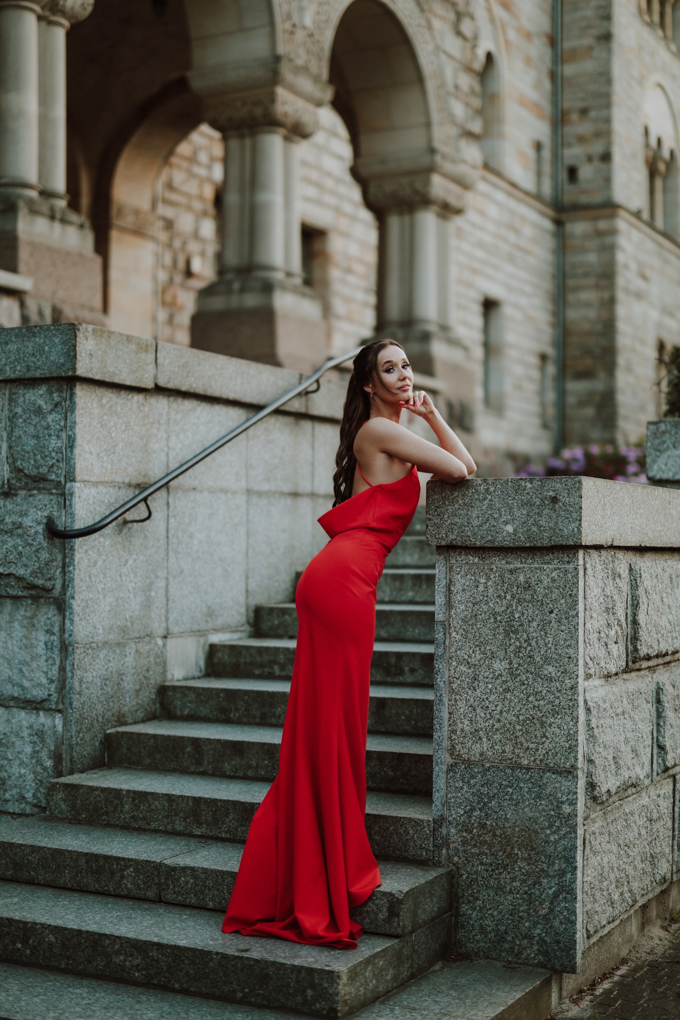 Kobieta w długiej czerwonej sukni pozuje na kamiennych schodach z balustradą, na tle zabytkowej architektury z kolumnami i łukami.