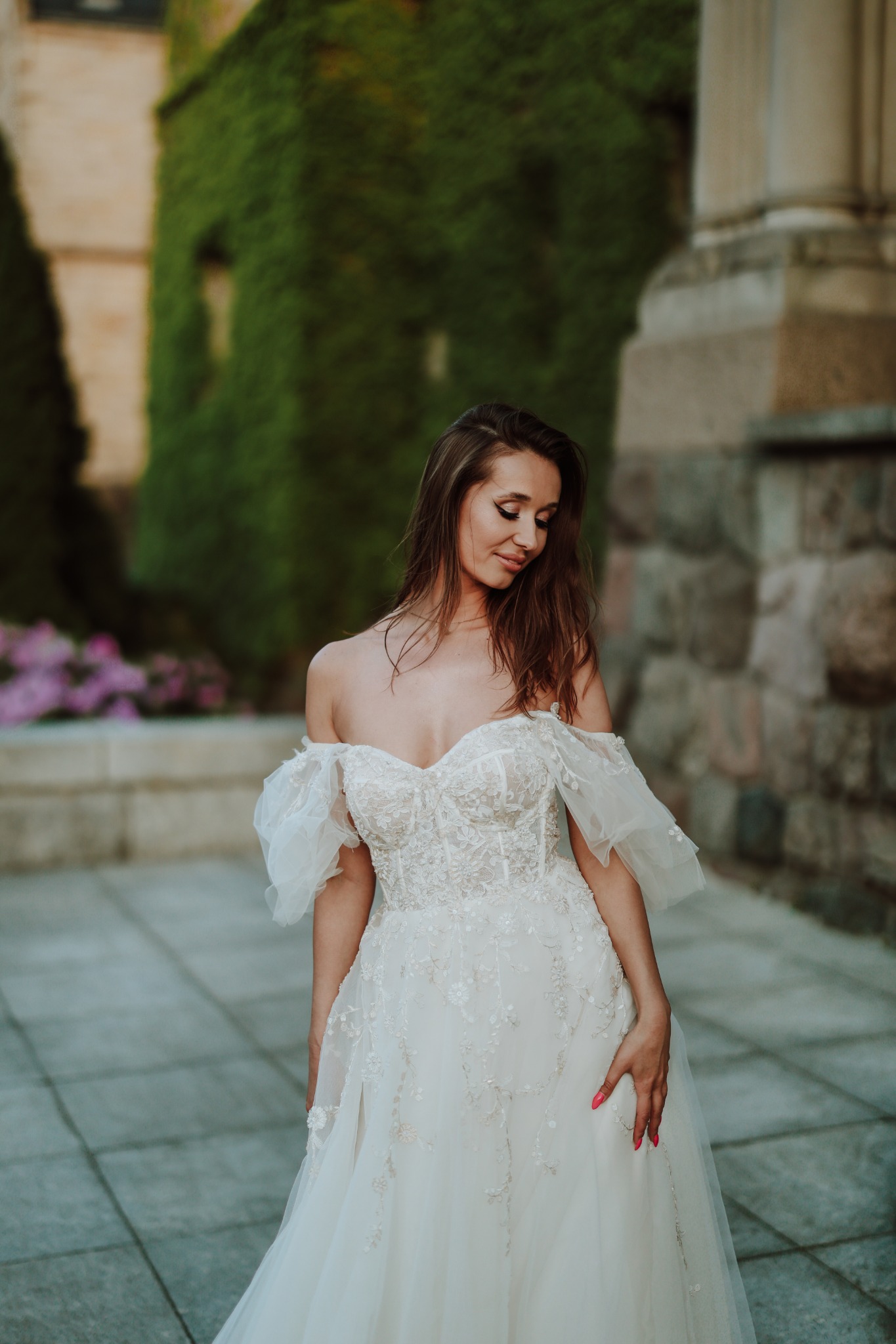 Panna młoda w koronkowej sukni ślubnej z odkrytymi ramionami, spoglądająca w dół, na tle kamiennej architektury porośniętej bluszczem.