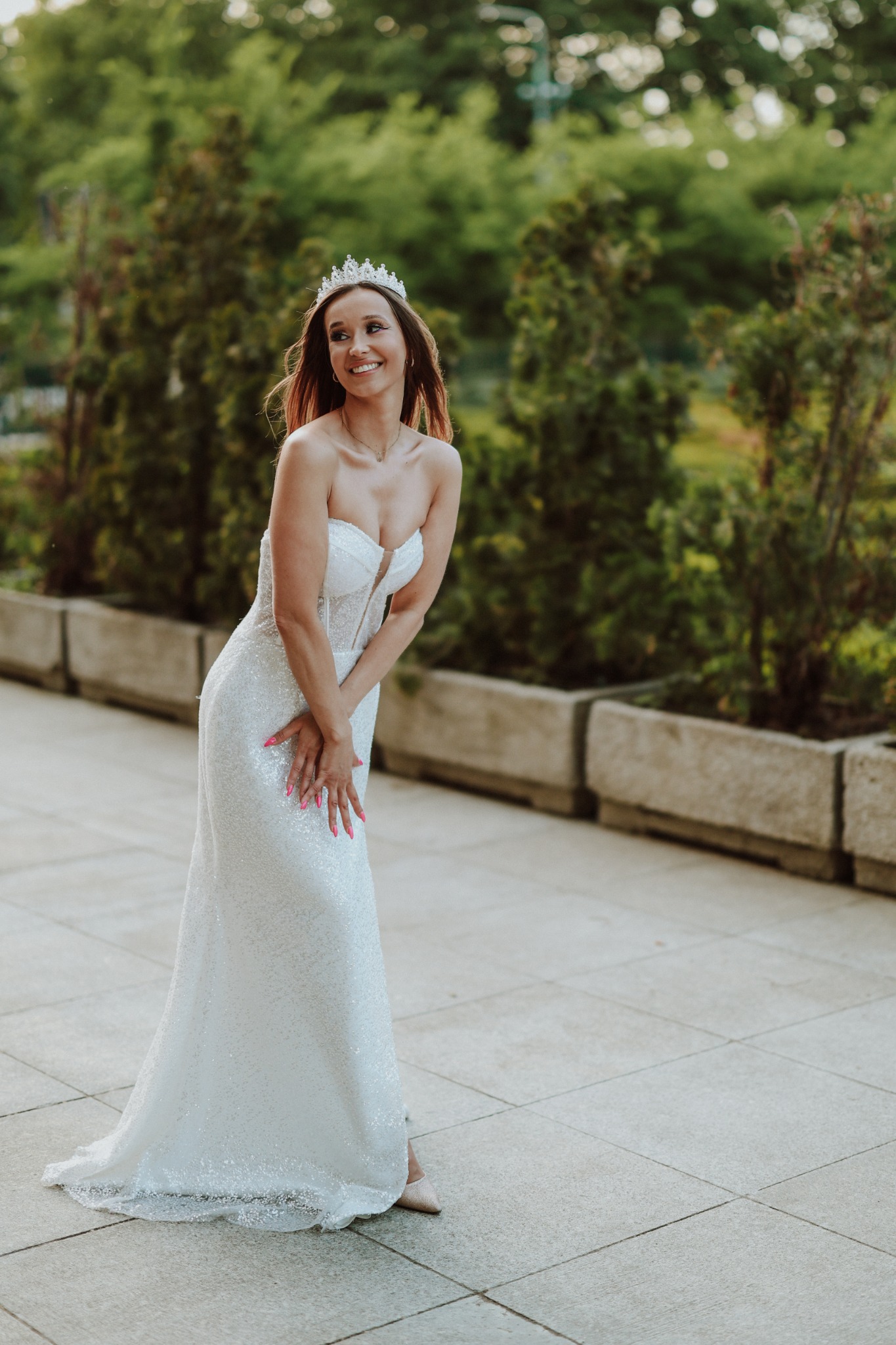 Uśmiechnięta kobieta w białej, błyszczącej sukni bez ramiączek i tiarze, pozuje na tle zieleni.